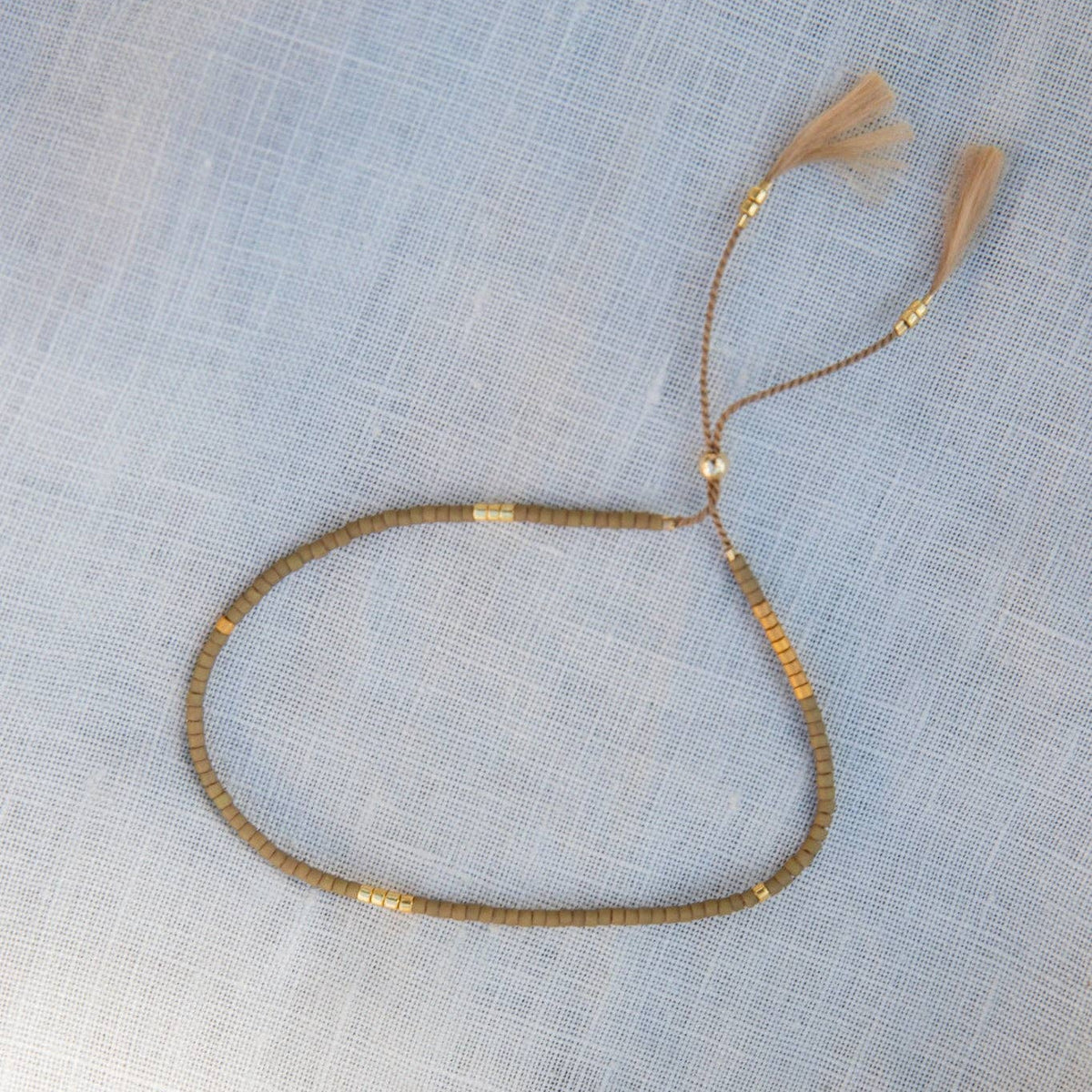 A beaded necklace with metallic gold-toned beads and light brown tassels, laid flat on a textured light blue fabric background.