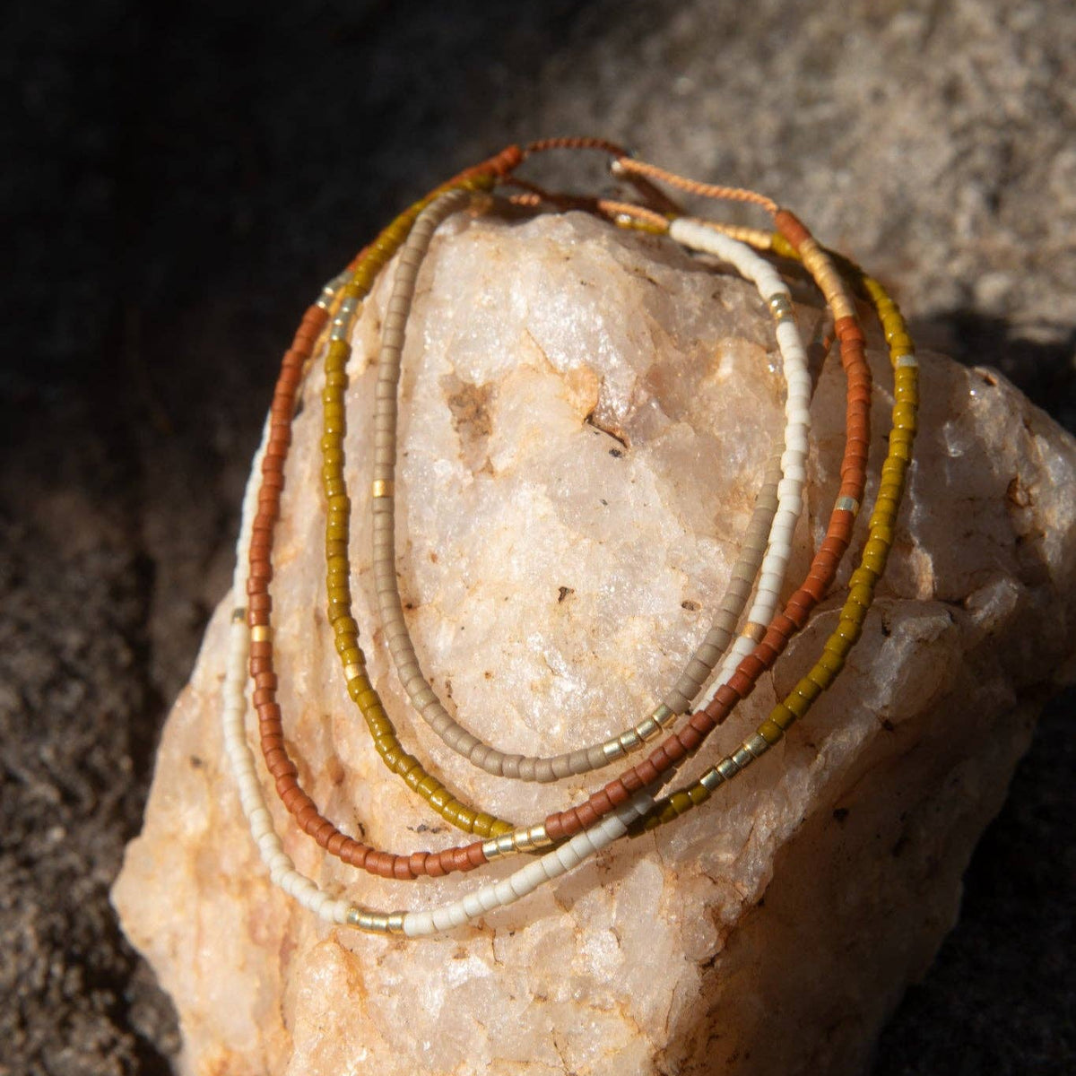 Three beaded bracelets in rust orange, olive green, and cream white, coiled on a pale pinkish stone against a dark textured rock background.