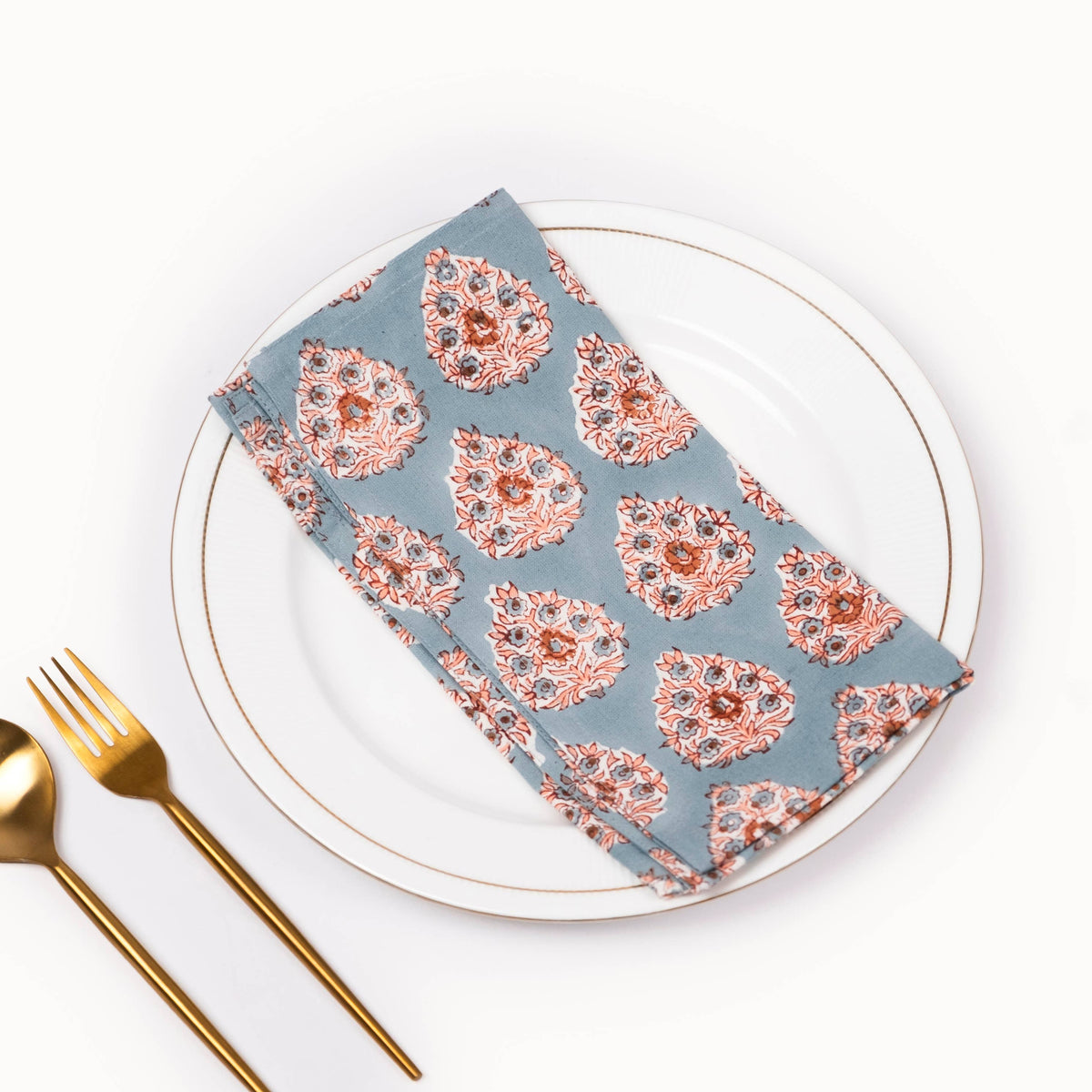 A folded patterned cloth napkin with a dusty blue-gray background and peach, pink, and white paisley-like motifs, styled on a white plate with gold cutlery on a white background.