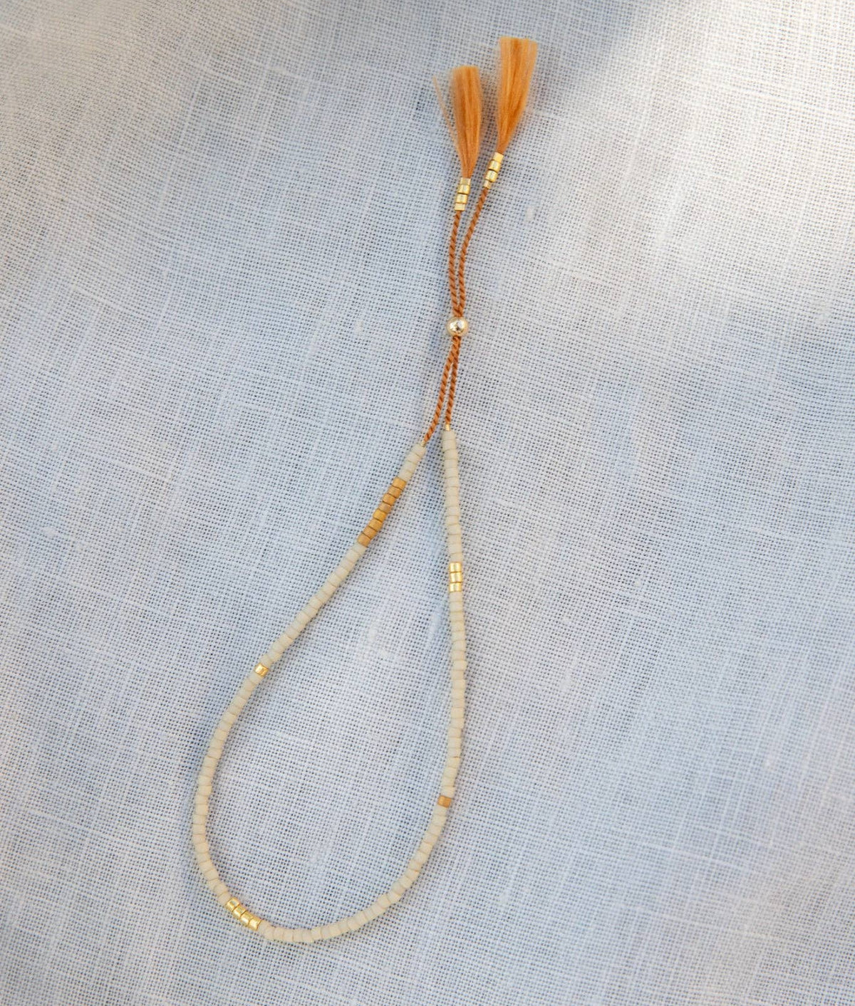 A beaded necklace with a loop of cream-colored beads, small gold spacer beads, and a brown cord tassel with orange tassels, laid flat on a light gray textured fabric background.
