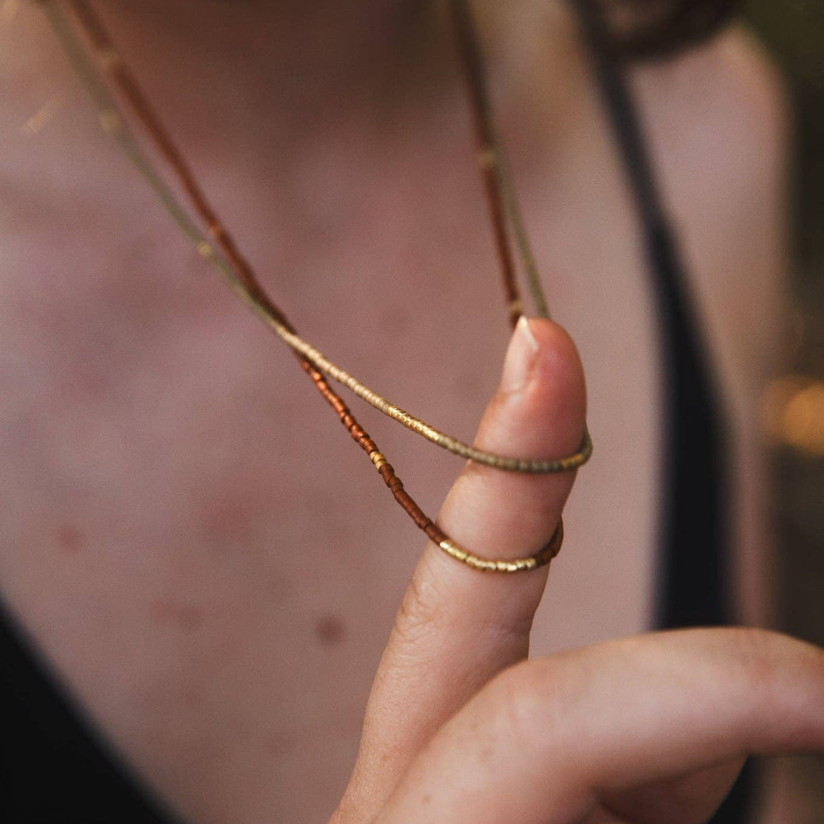 A close-up lifestyle shot of a person wearing a delicate double-layered necklace with one layer in yellow gold and the other in a textured rose gold or copper tone, draped over their chest and held by their finger.