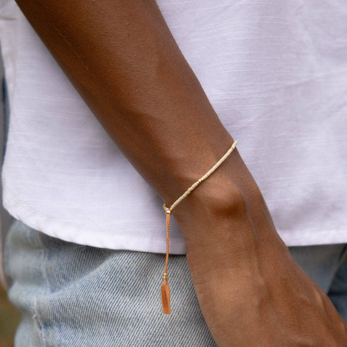 A close-up of a person's wrist wearing a delicate gold chain bracelet with a small dangling tassel, styled with a white t-shirt and light blue jeans.