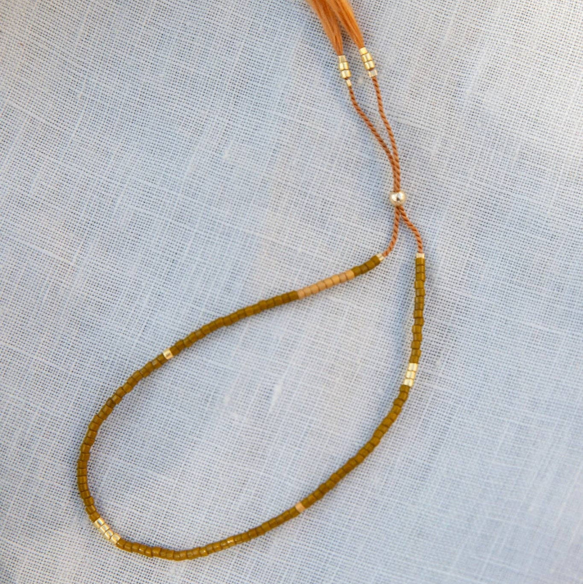 A beaded bracelet with olive green and light brown beads, accented with small gold spacer beads, featuring a matching light brown tassel, laid out on a textured light blue fabric background.