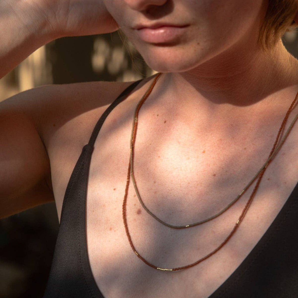 A close-up lifestyle shot of a woman wearing a double-strand bronze chain necklace with a small central disc, paired with a black tank top.