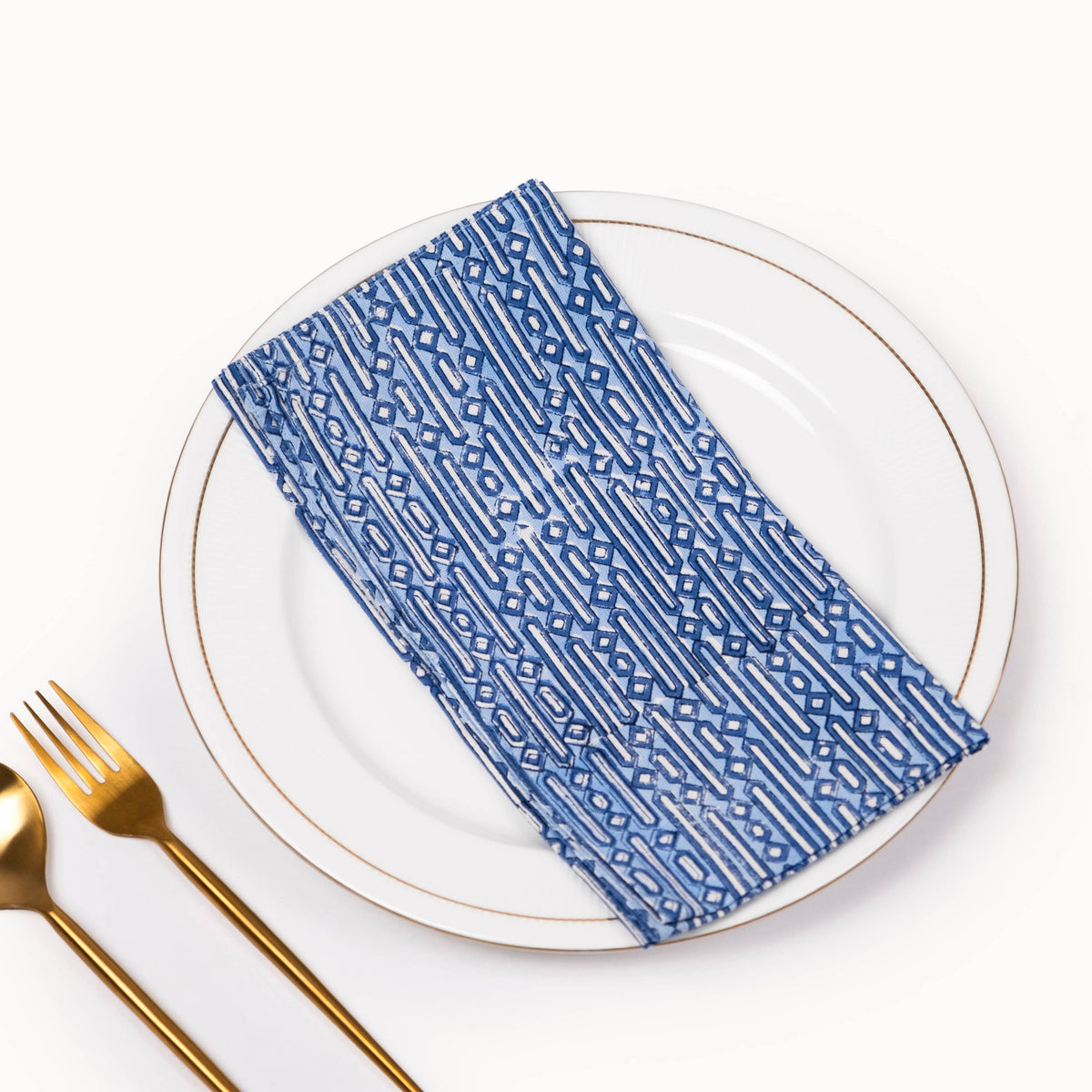 A blue and white patterned cloth napkin folded on a white plate with gold trim, styled with gold-toned cutlery on a clean white background.