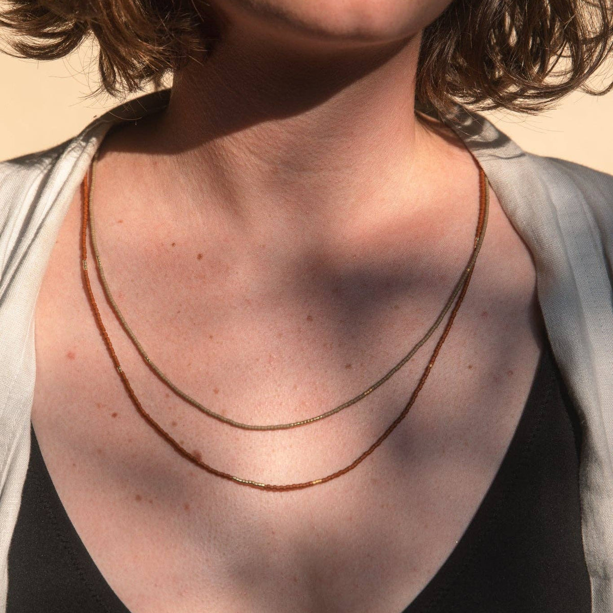 A close-up of a person wearing a delicate layered necklace with small amber-like beads over a black top and light gray cardigan, against a warm beige background.