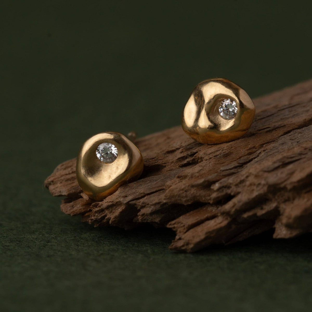 A pair of gold stud earrings with a small clear gemstone, resting on a piece of textured dark wood against a dark green background.