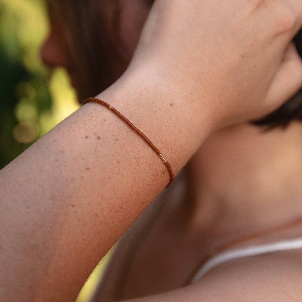 A close-up lifestyle shot of a person wearing a thin brown cord bracelet with a small gold bead accent on their wrist, set against a soft-focus outdoor background.