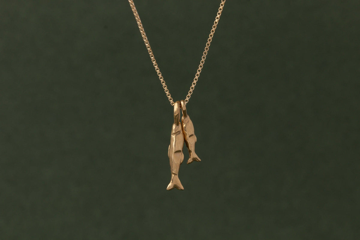 A gold pendant necklace featuring two small fish hanging from a delicate gold chain, set against a dark olive green background.