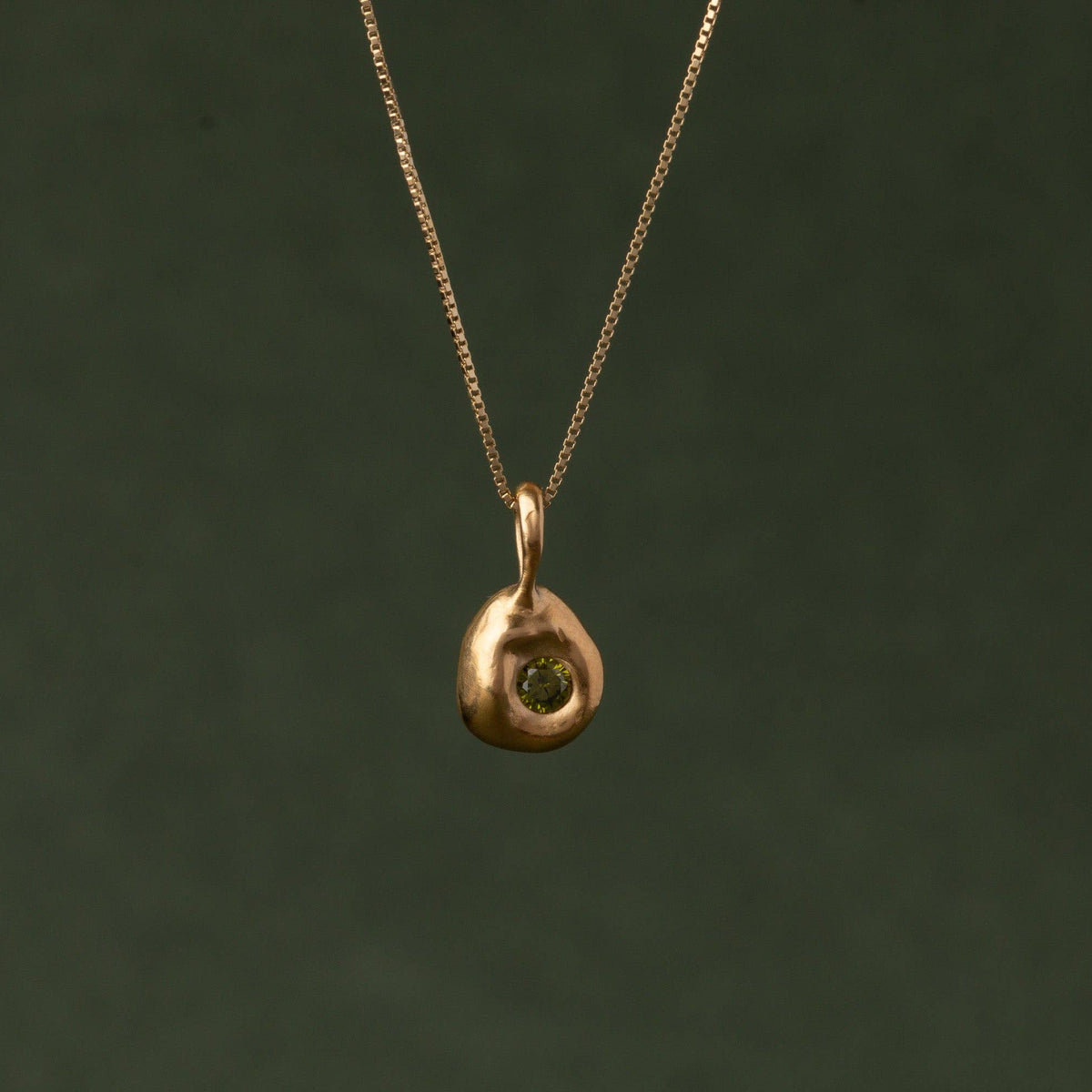 A gold necklace with a small, organic-shaped gold pendant featuring a central green gemstone, suspended against a dark olive green background.
