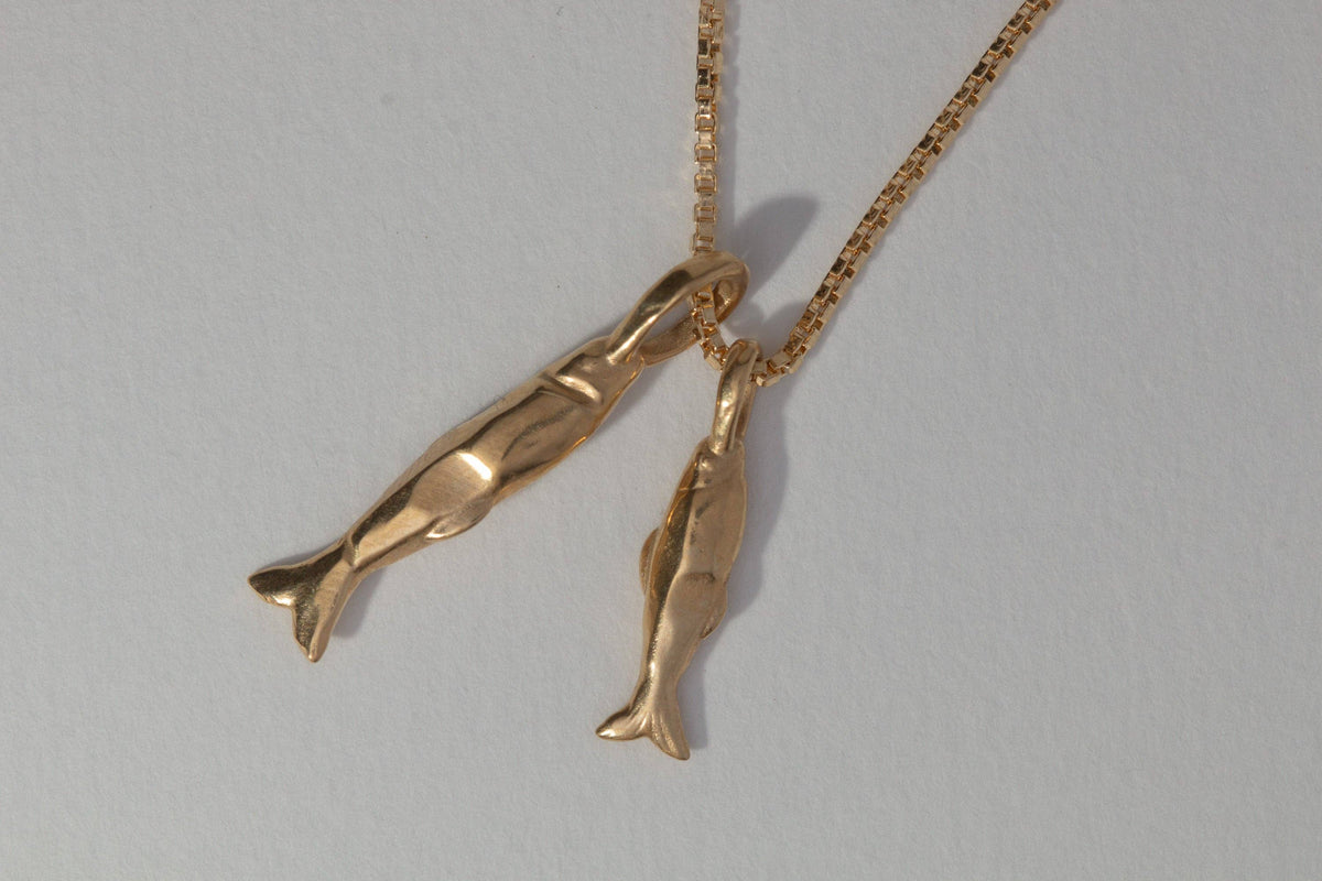 A gold necklace with two small fish or dolphin-shaped pendants hanging from a delicate chain, photographed against a plain light gray background.