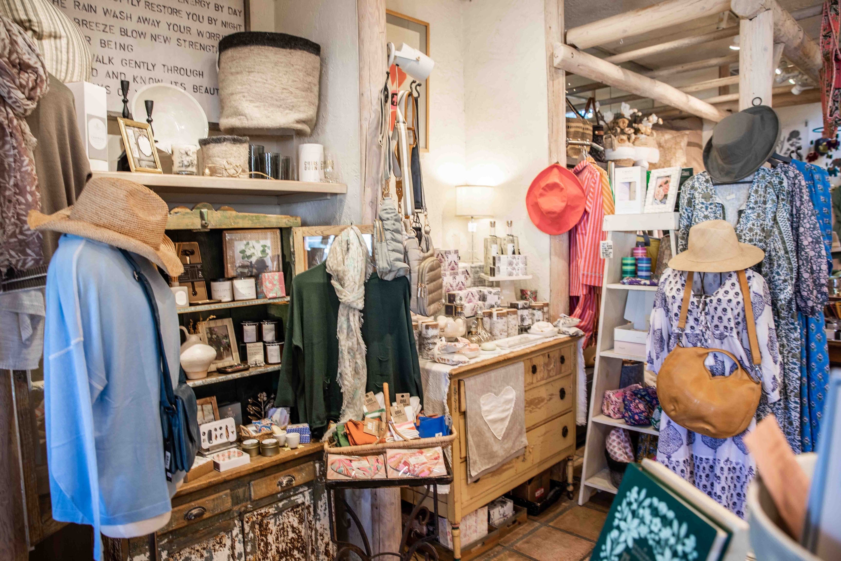 A cozy, rustic boutique interior filled with vintage clothing, hats, and home decor. A light blue cardigan and a tan wide-brim hat are displayed on a wooden stand on the left, while a blue patterned dress and a brown leather shoulder bag hang on a mannequin on the right.
