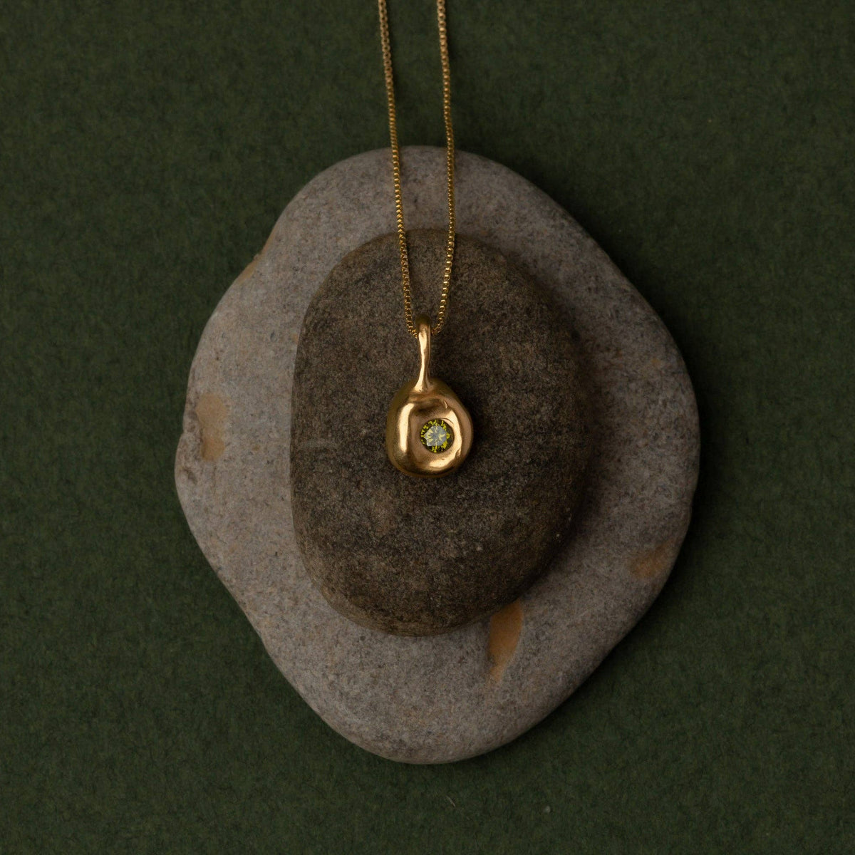 A gold pendant necklace with a small oval gemstone, displayed on a dark gray stone against a dark green background.