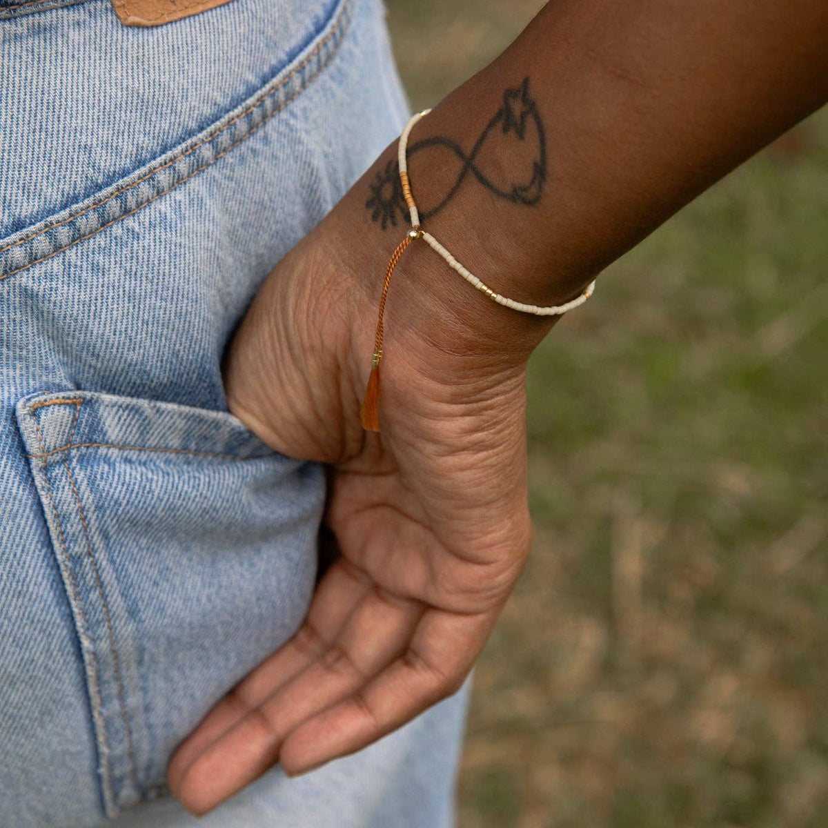 A close-up of a person's hand in a jean pocket wearing a delicate gold chain bracelet with a small amber bead and a black infinity symbol tattoo on the wrist.