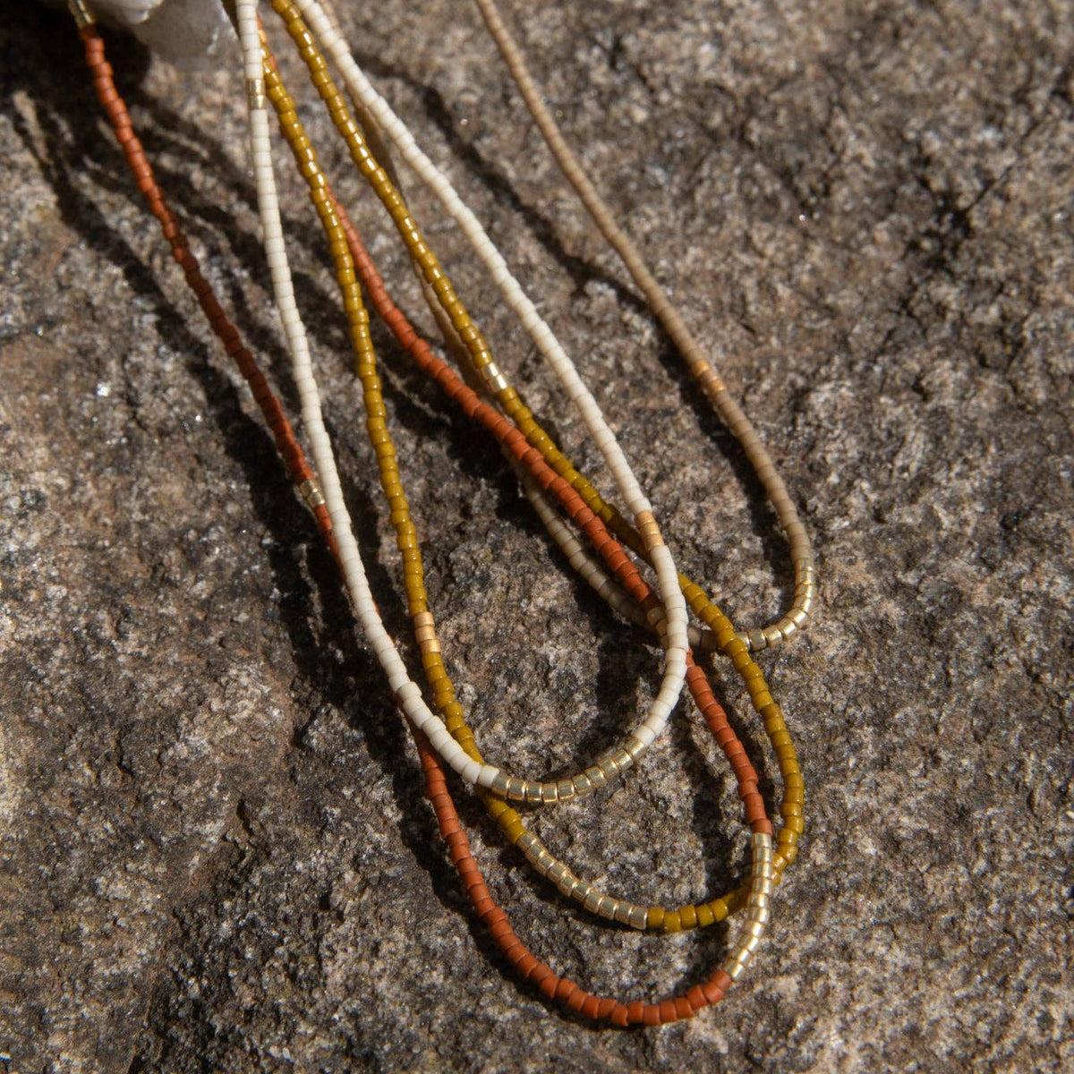Close-up of several thin cords or threads in cream, tan, orange, and metallic gold resting on a rough gray stone surface.