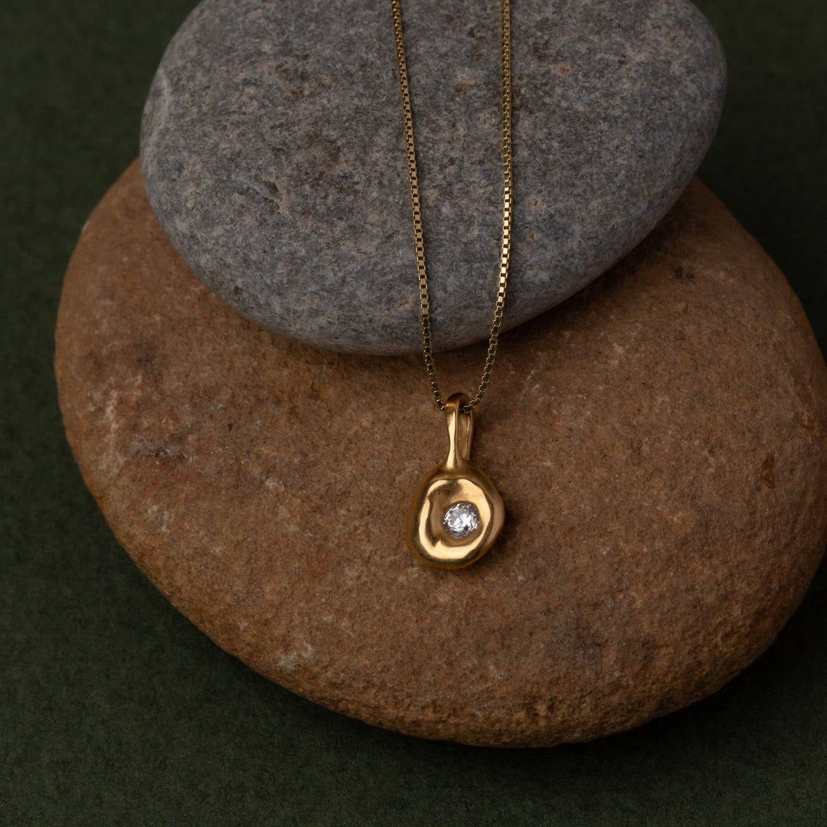 A gold pendant necklace with a small clear gemstone, displayed on a brown and gray stone stack against a dark green background.
