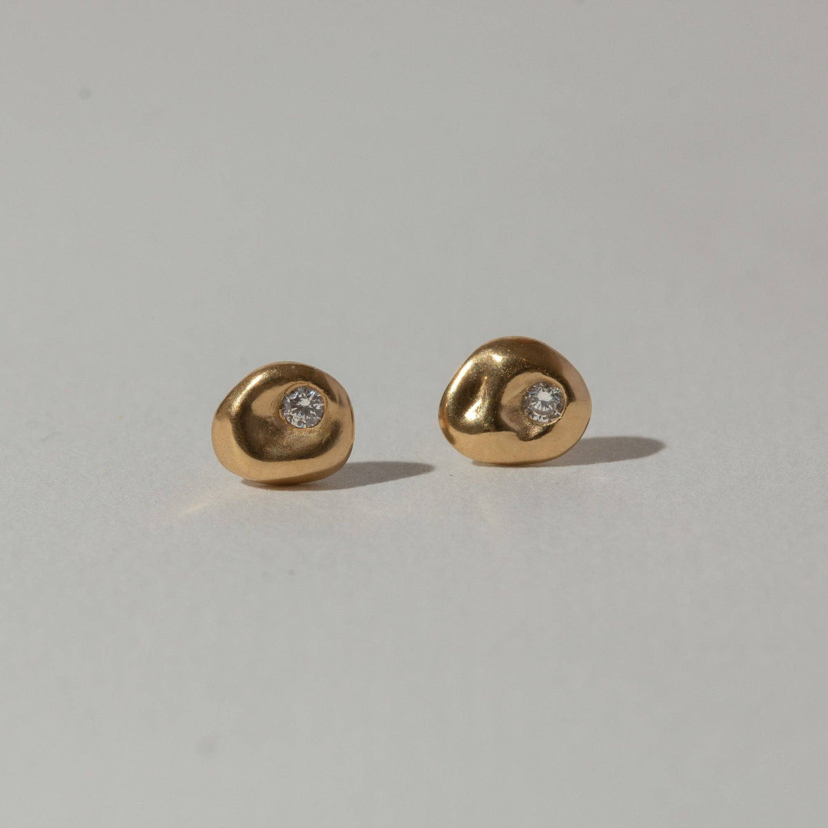 A pair of gold stud earrings with a small clear gemstone set in an organic, irregular shape, displayed on a light gray background.