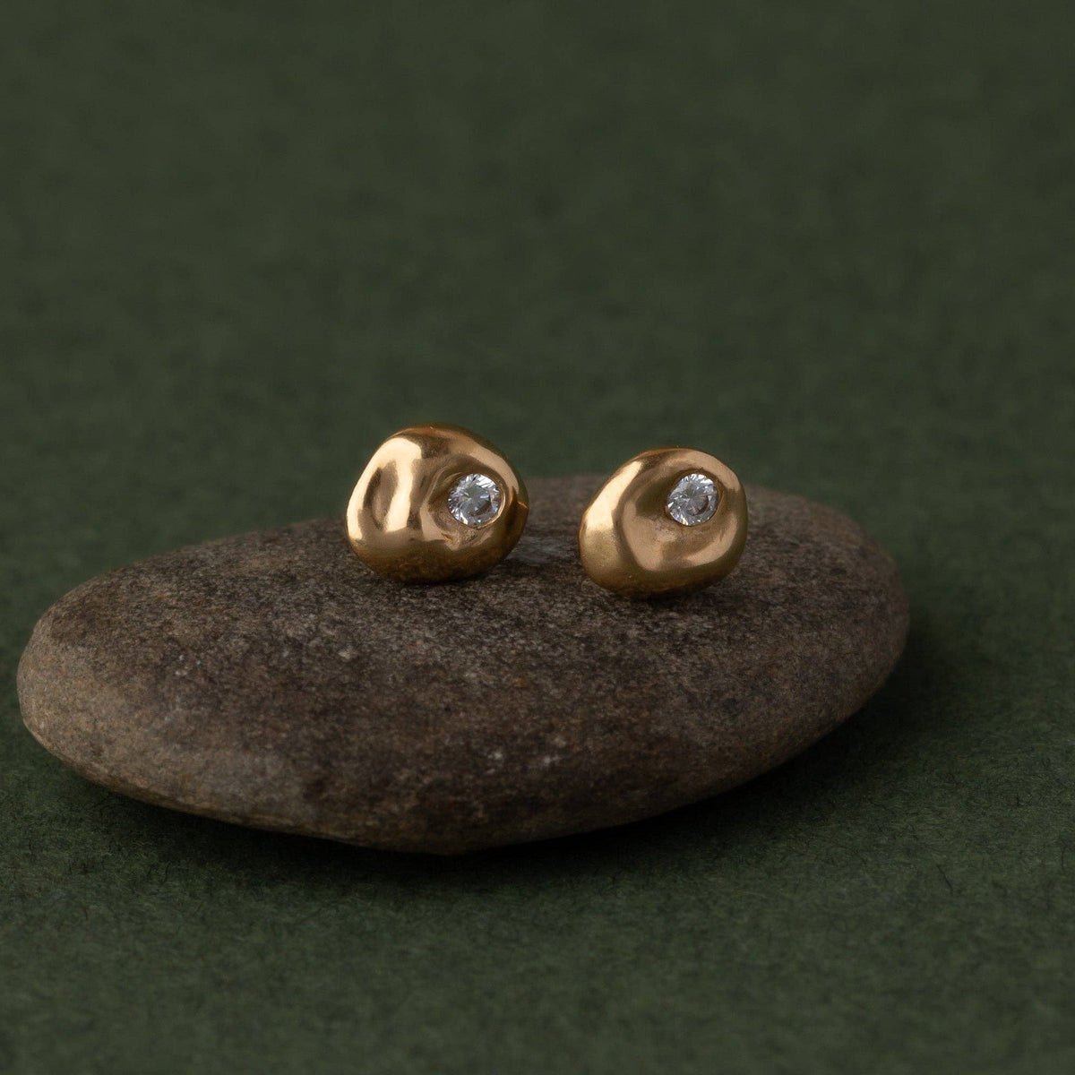 A pair of small gold stud earrings with a tiny clear gemstone, displayed on a dark stone against a dark green background.