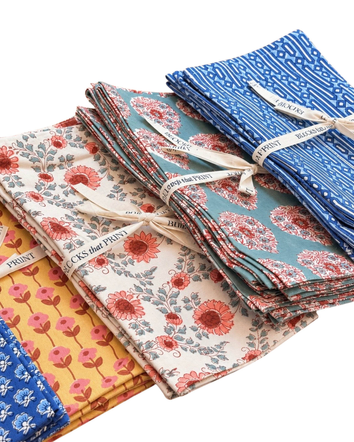 A stack of six folded cotton fabric napkins in various floral and geometric patterns, tied with white paper bands. The fabrics feature blue, teal, yellow, and cream colors with red, pink, and white motifs, set against a plain white background.