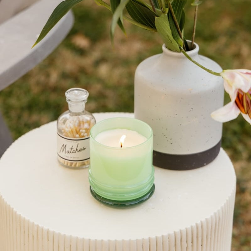 Lit candle in green jar on a patio side table with a vase and matches