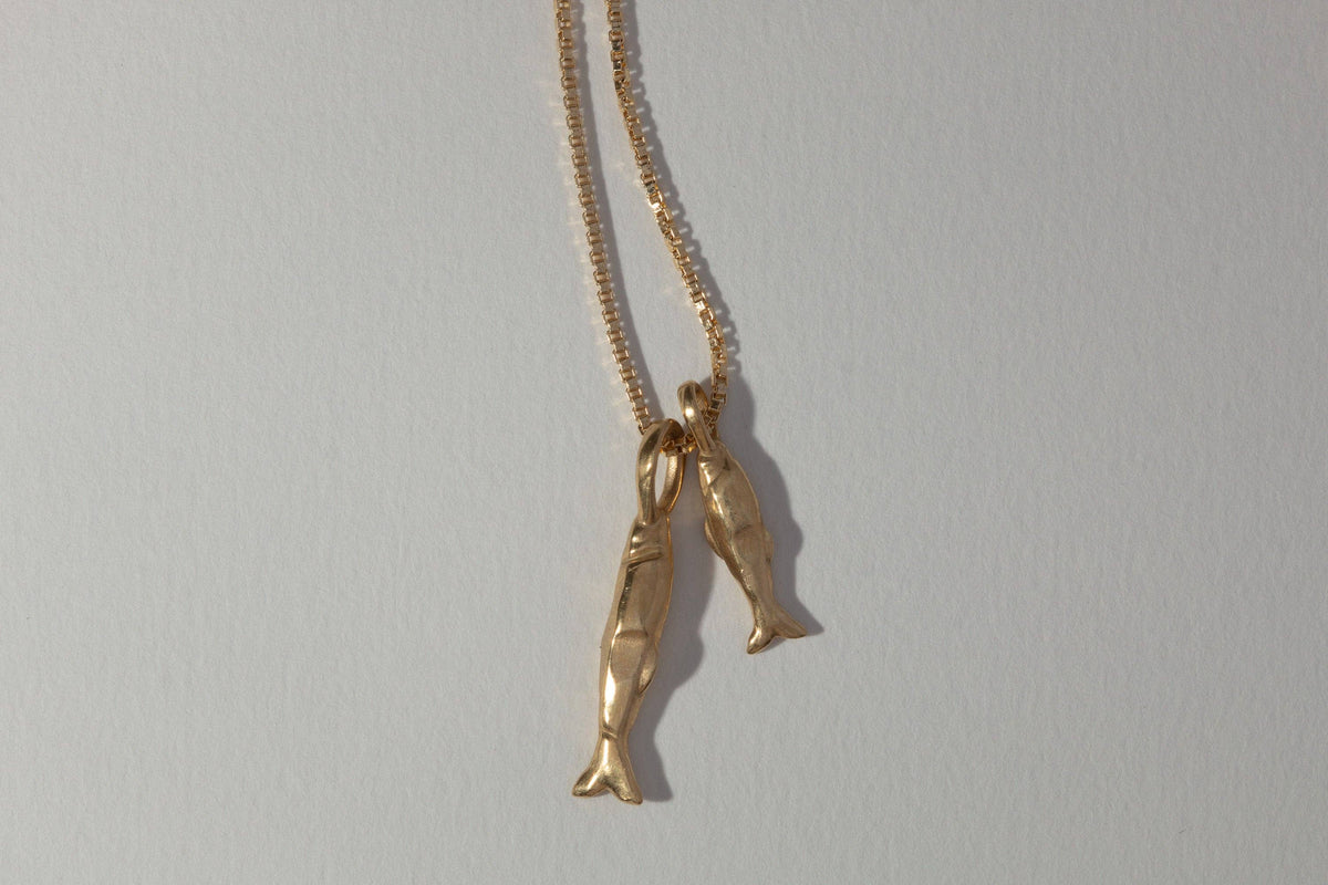 A gold chain necklace featuring two dangling fish charms, photographed against a plain light gray background.