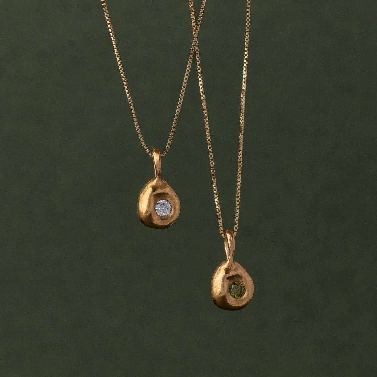 Two gold teardrop-shaped pendant necklaces with small clear and green gemstones, hanging against a dark olive green background.