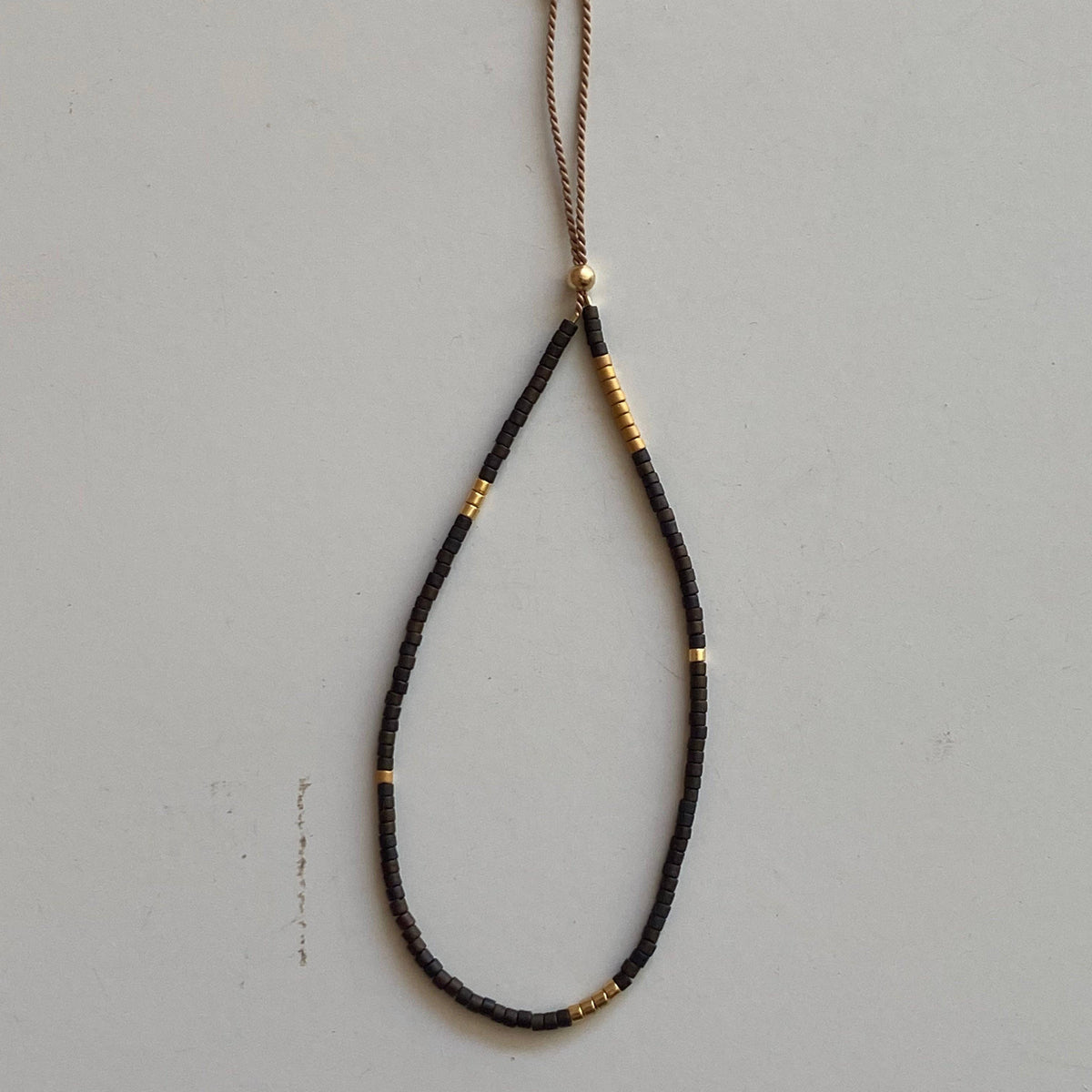 A black braided cord bracelet with small gold metallic beads and a light tan tassel, displayed on a neutral light gray background.