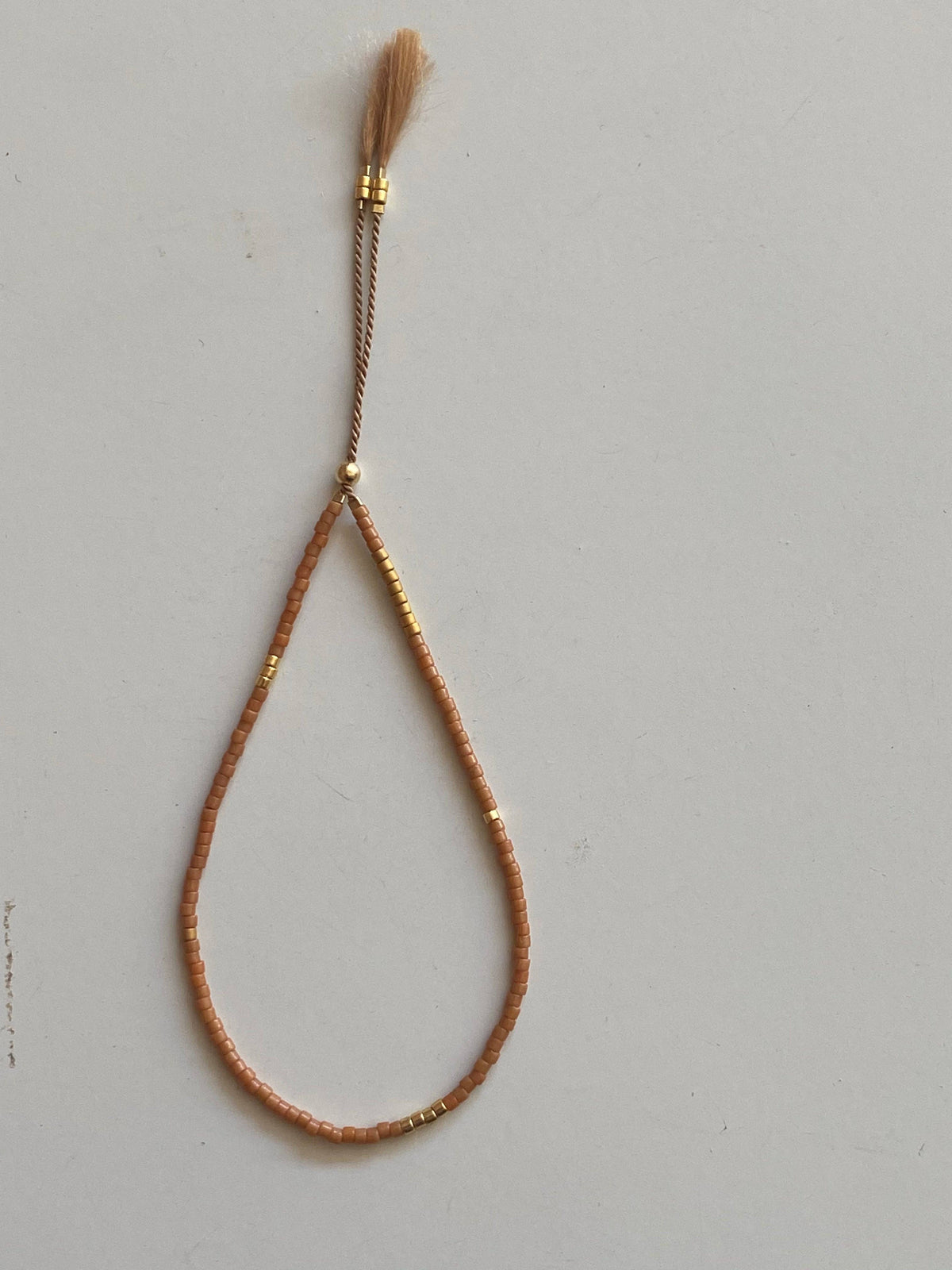 A delicate teardrop-shaped beaded necklace in a warm peachy-brown color with small gold metallic accents, featuring a matching tassel on a thin cord, displayed against a light gray background.