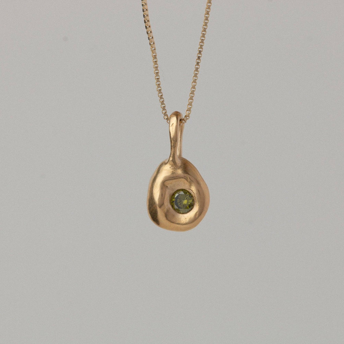 A gold pendant necklace with a small, organic-shaped gold drop featuring a central green gemstone, suspended against a plain light gray background.