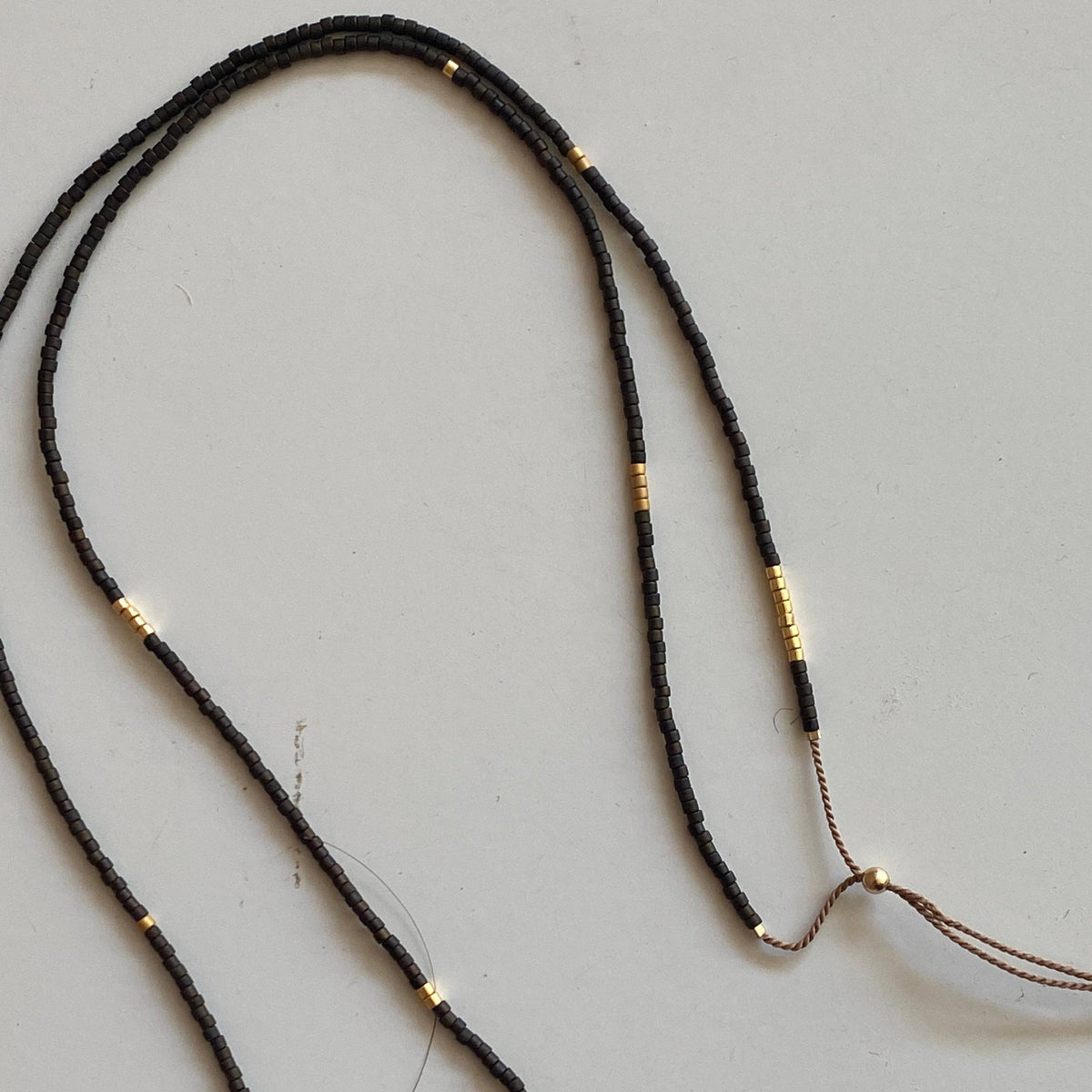 A black braided cord necklace with small gold spacer beads and a gold-toned clasp, laid out on a light gray surface.
