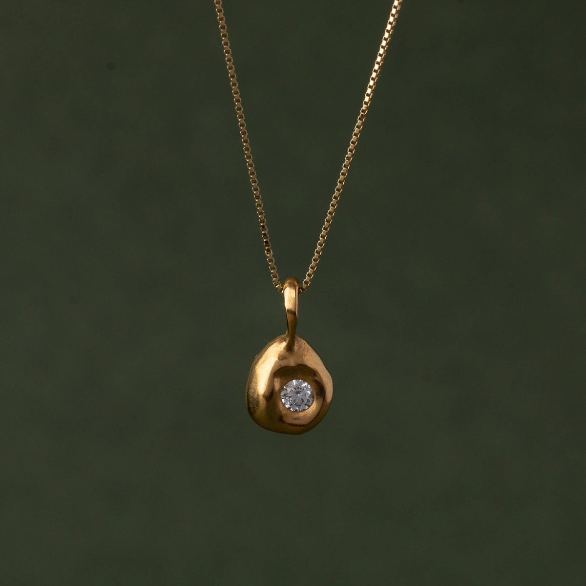 A gold teardrop-shaped pendant necklace with a small clear gemstone in the center, hanging against a dark olive green background.