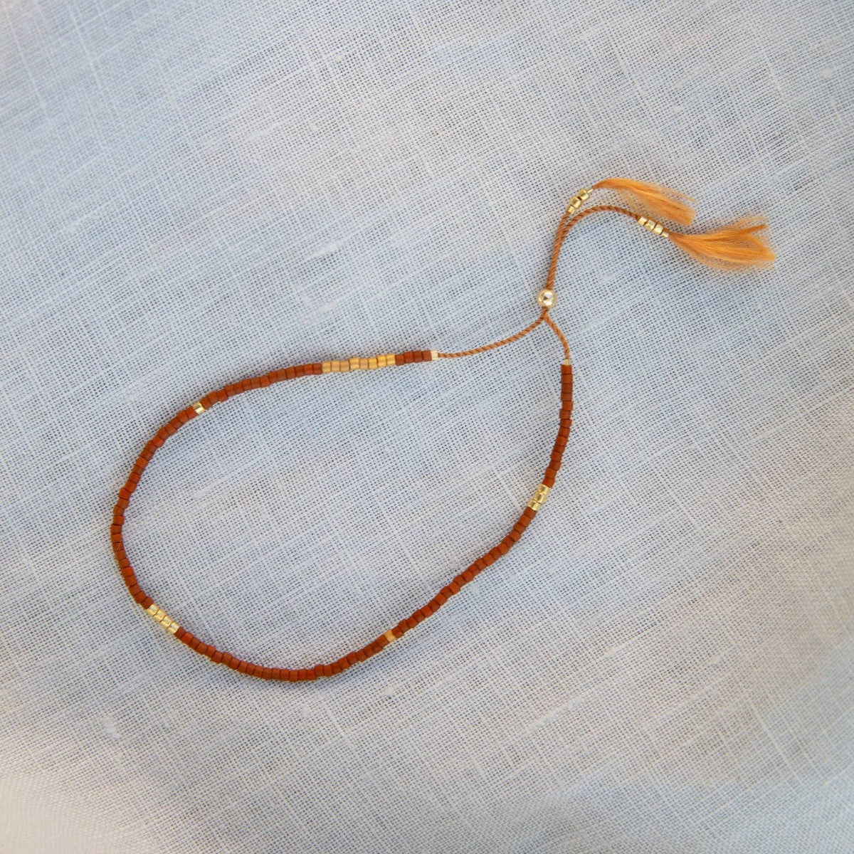 A delicate beaded bracelet with a reddish-brown cord, small gold spacer beads, and a matching peach tassel, laid flat on a light blue textured fabric background.