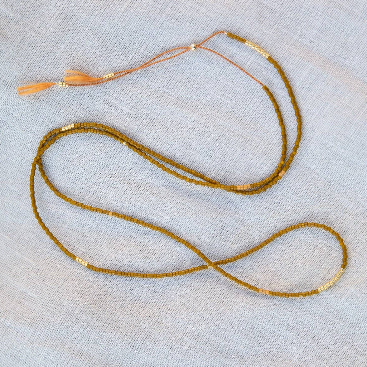 A delicate gold beaded necklace with a small orange tassel, laid out on a light beige textured fabric background.