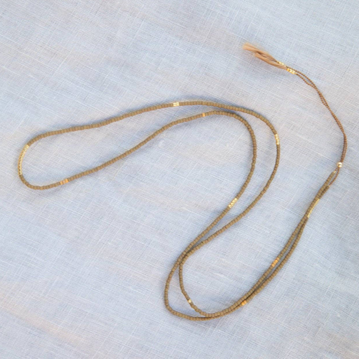 A delicate beaded necklace with small metallic beads, laid out on a light gray textured fabric background.