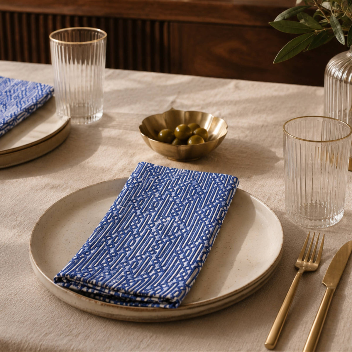 A styled dining table setting featuring blue patterned linen napkins on beige ceramic plates, accompanied by clear ribbed glass tumblers, gold-toned flatware, and a small gold bowl of olives, with a warm neutral background and soft shadows.
