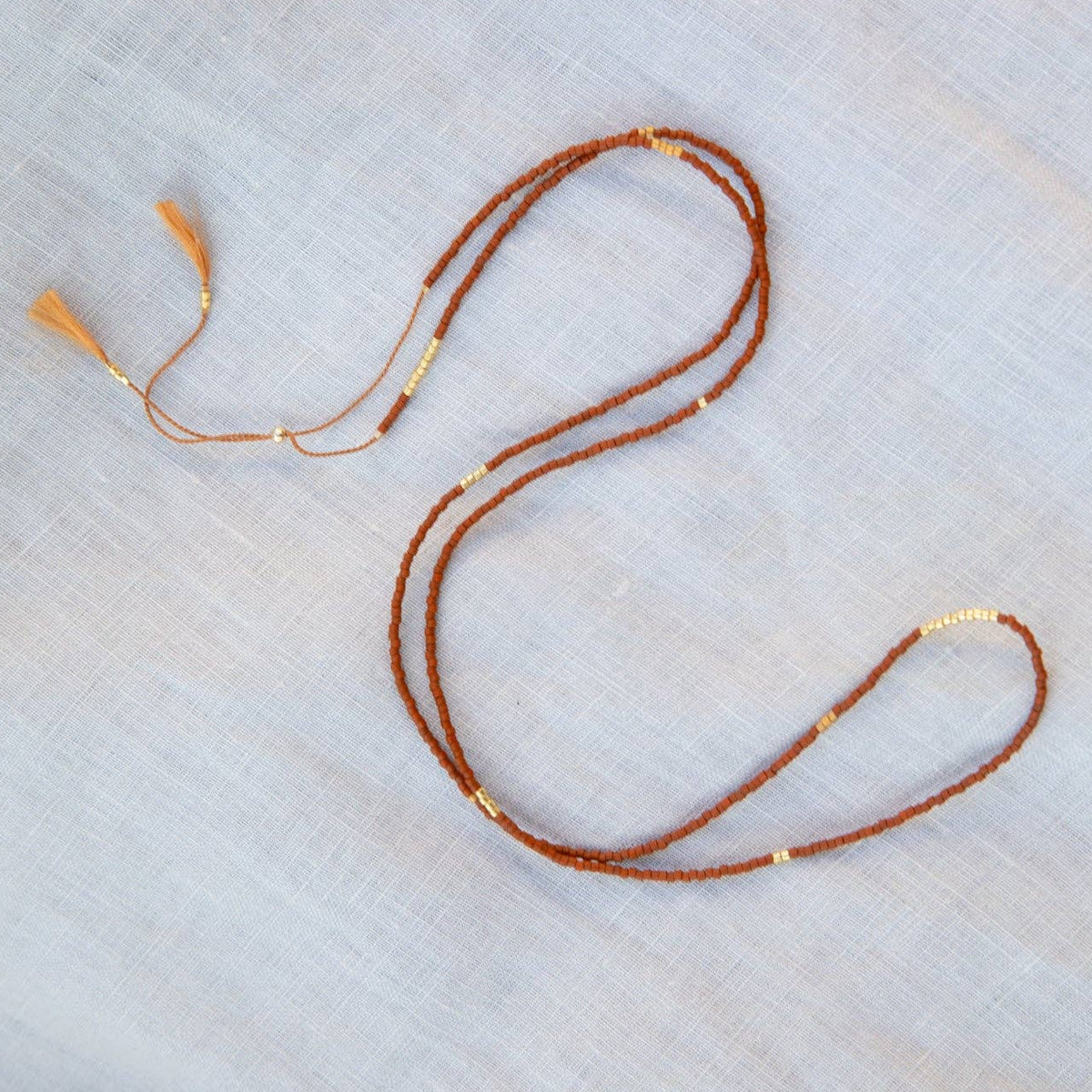 A delicate single-strand necklace with small brown beads and tiny gold spacer beads, laid out on a light gray textured fabric background.