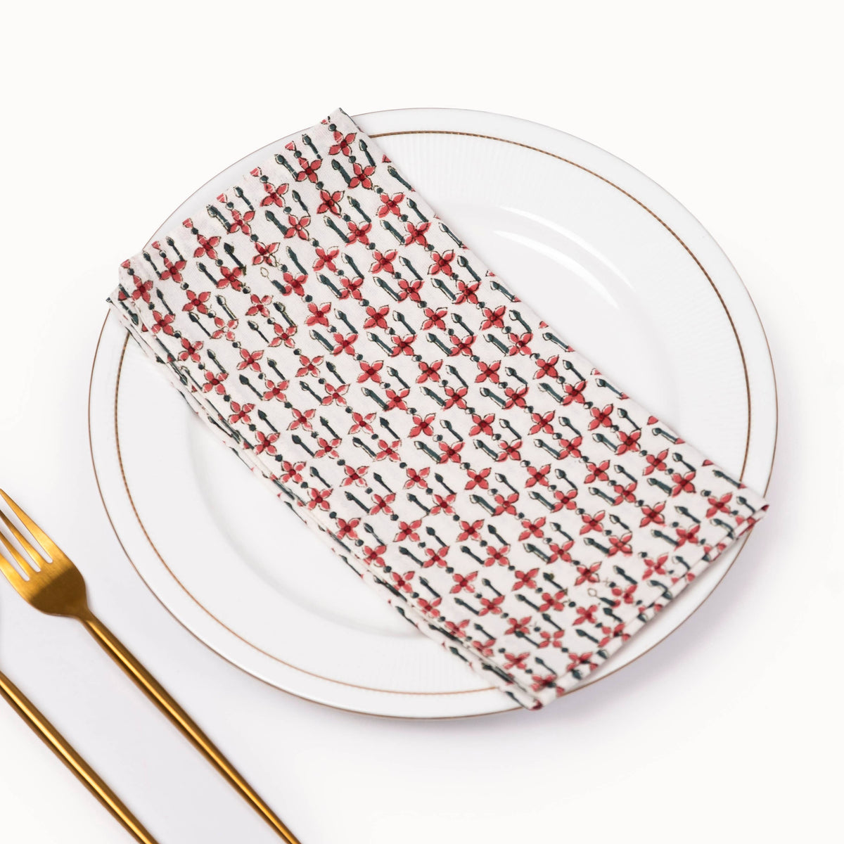 A folded white napkin with a repeating pattern of red and black floral silhouettes, styled on a white ceramic plate with gold trim, alongside gold-toned cutlery on a white background.