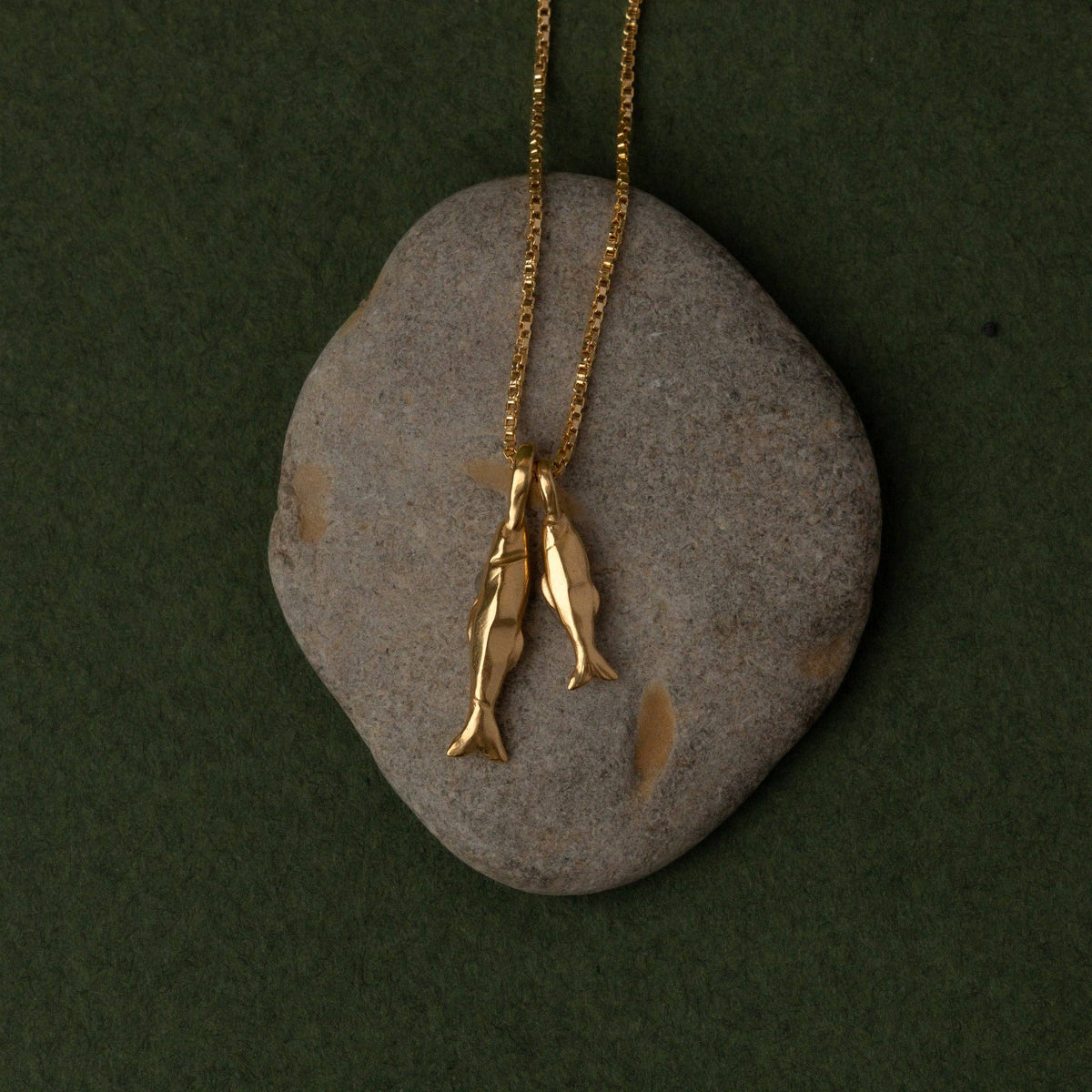 A gold necklace with two small fish pendants hanging from a gold chain, displayed on a smooth gray stone against a dark green background.