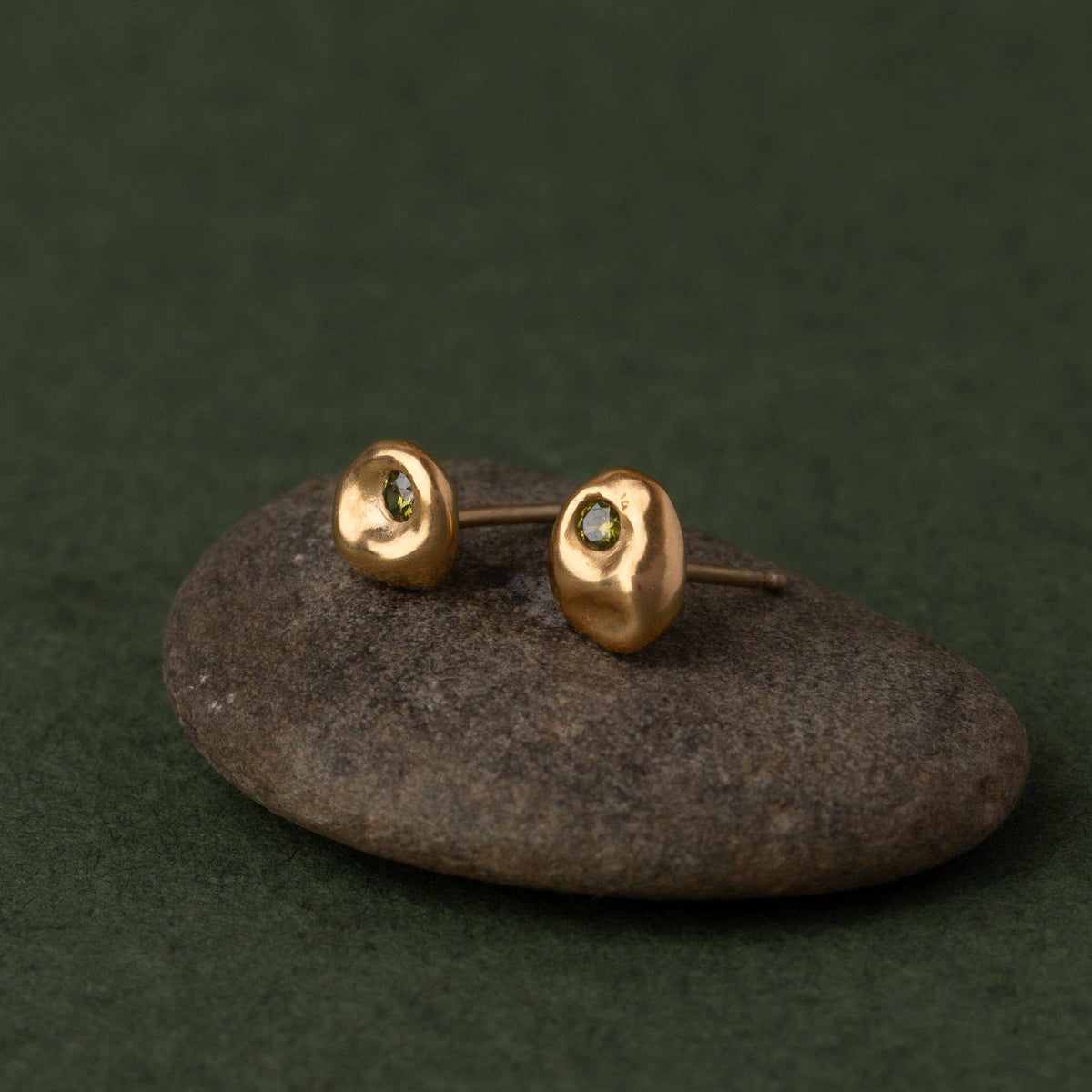 A pair of small gold stud earrings with oval textured tops and tiny dark green gemstones, displayed on a smooth dark stone against a dark green background.