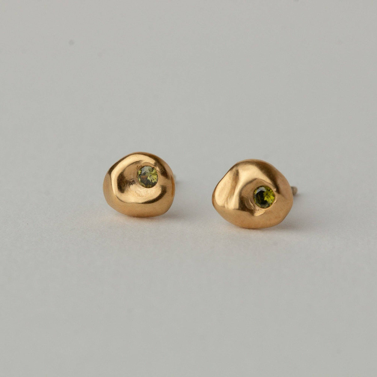 A pair of gold stud earrings with organic, rounded shapes and a small green gemstone set in the center, displayed on a neutral light gray background.