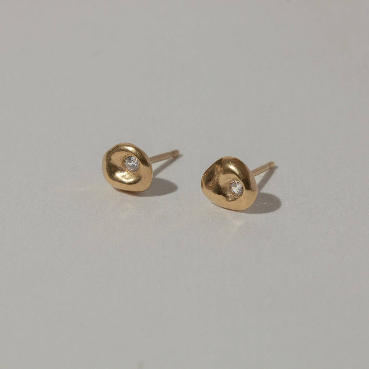 A pair of small gold stud earrings with a bezel-set clear gemstone, displayed on a neutral light gray surface.