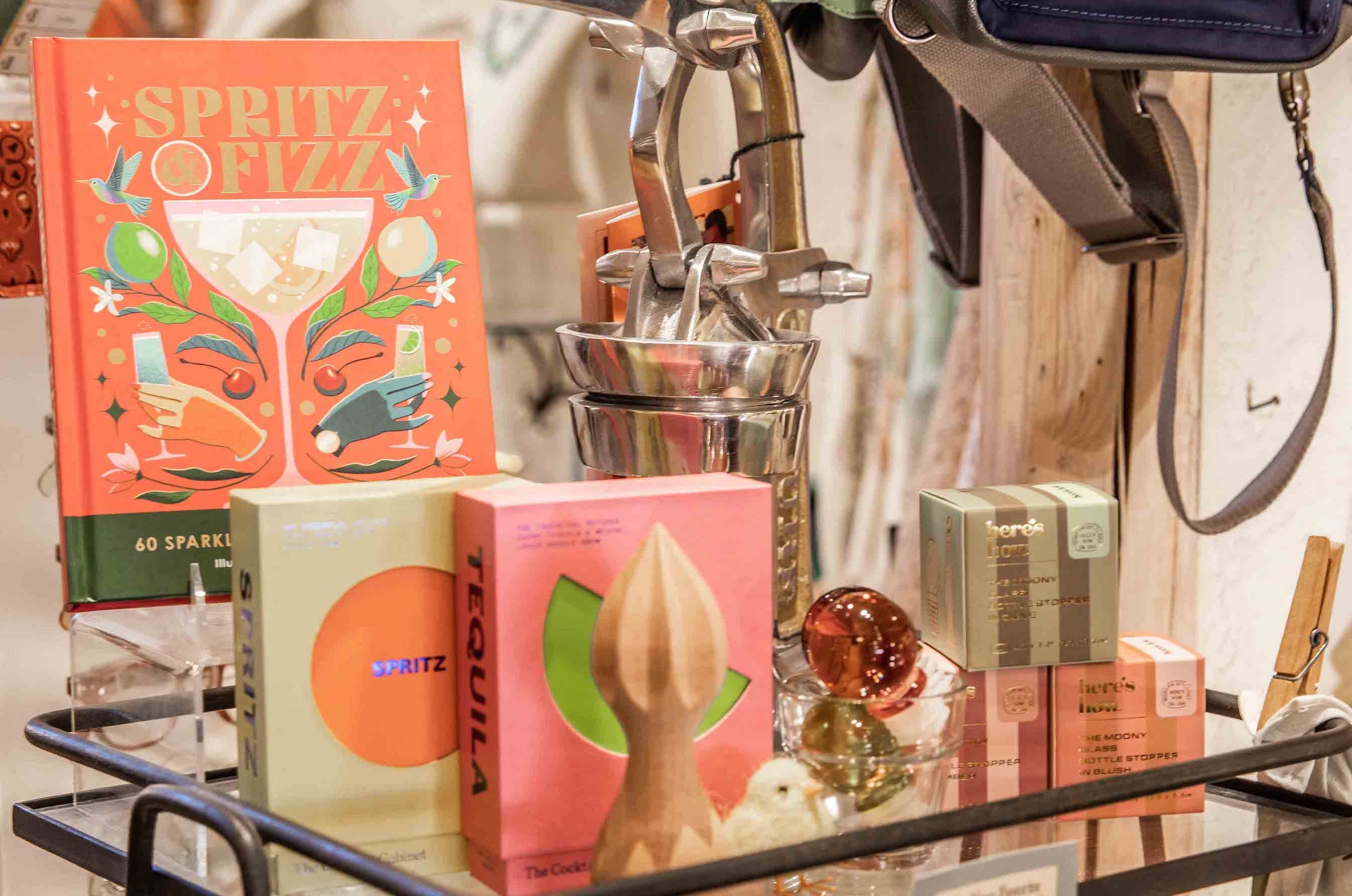 A styled retail display featuring a prominent orange book titled 'SPRITZ OF FIZZ' with cocktail illustrations, alongside several colorful books on a glass shelf next to a wooden juice reamer.