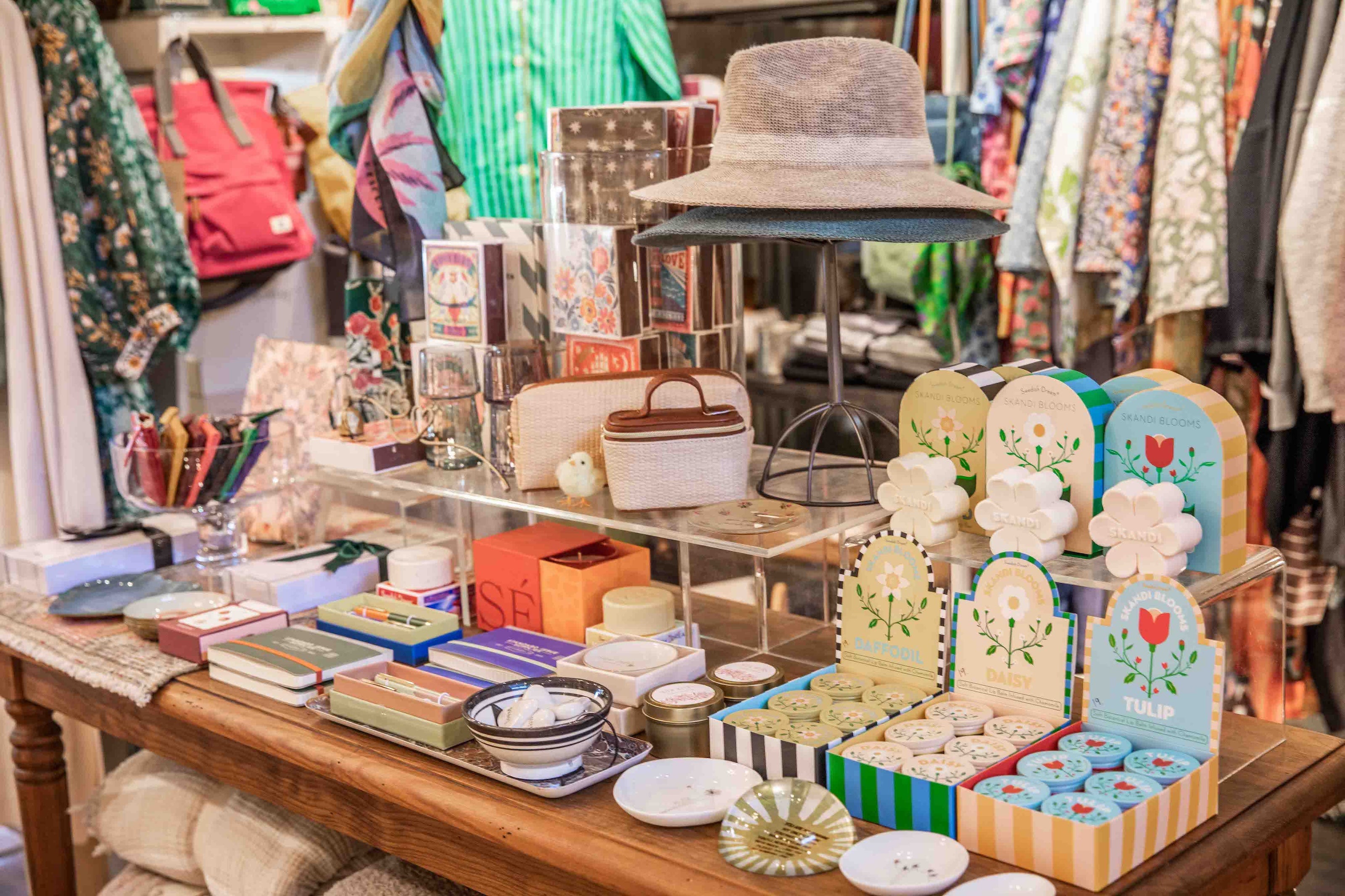 A retail display table filled with assorted stationery, small boxes, decorative cards, and a woven hat, photographed inside a shop with clothing racks in the background.