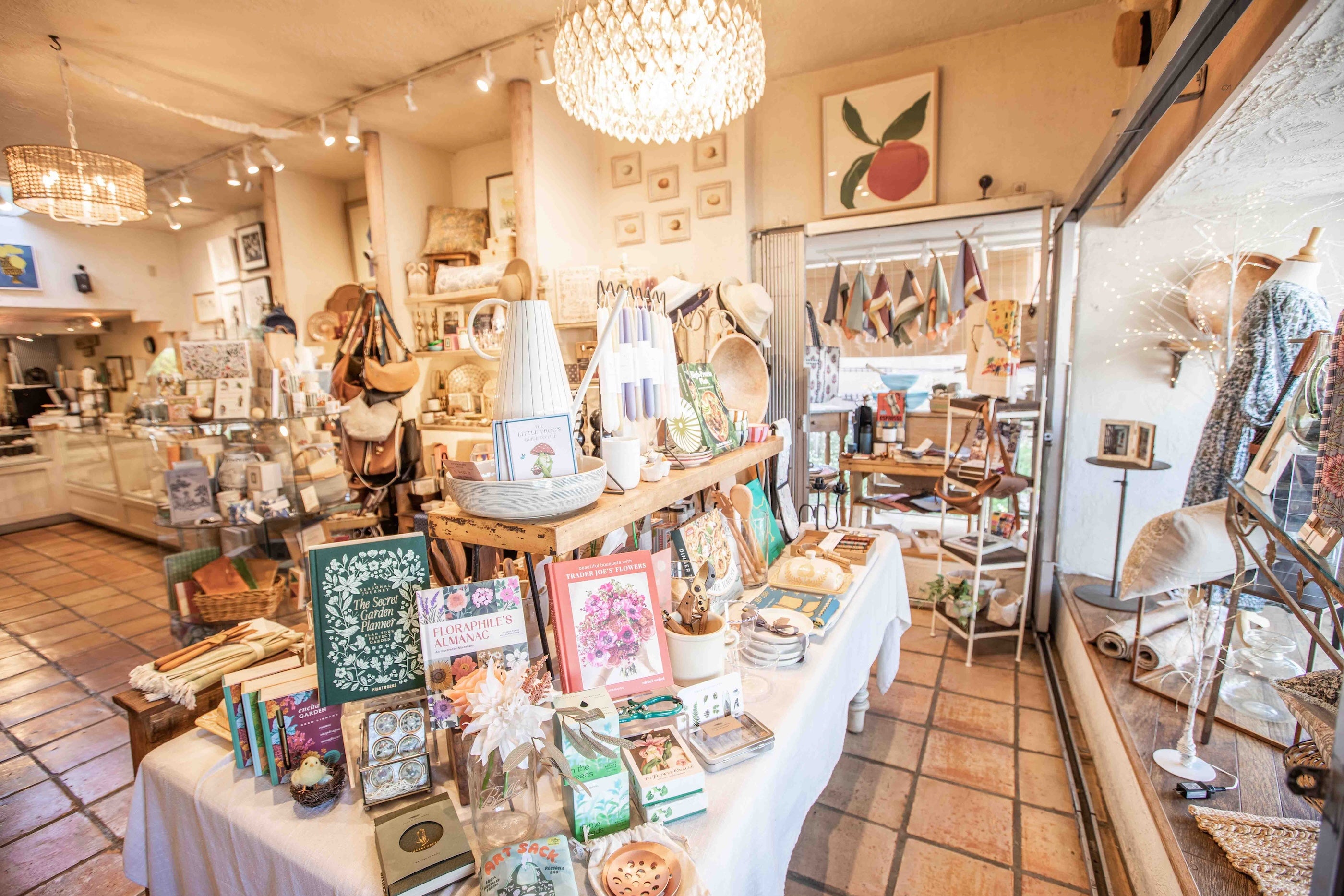 A cozy, rustic gift shop interior filled with handmade crafts, home decor, and books. Tables covered in white cloths display an assortment of items including framed art, small vases, candles, and packaged goods under warm, glowing overhead lights.