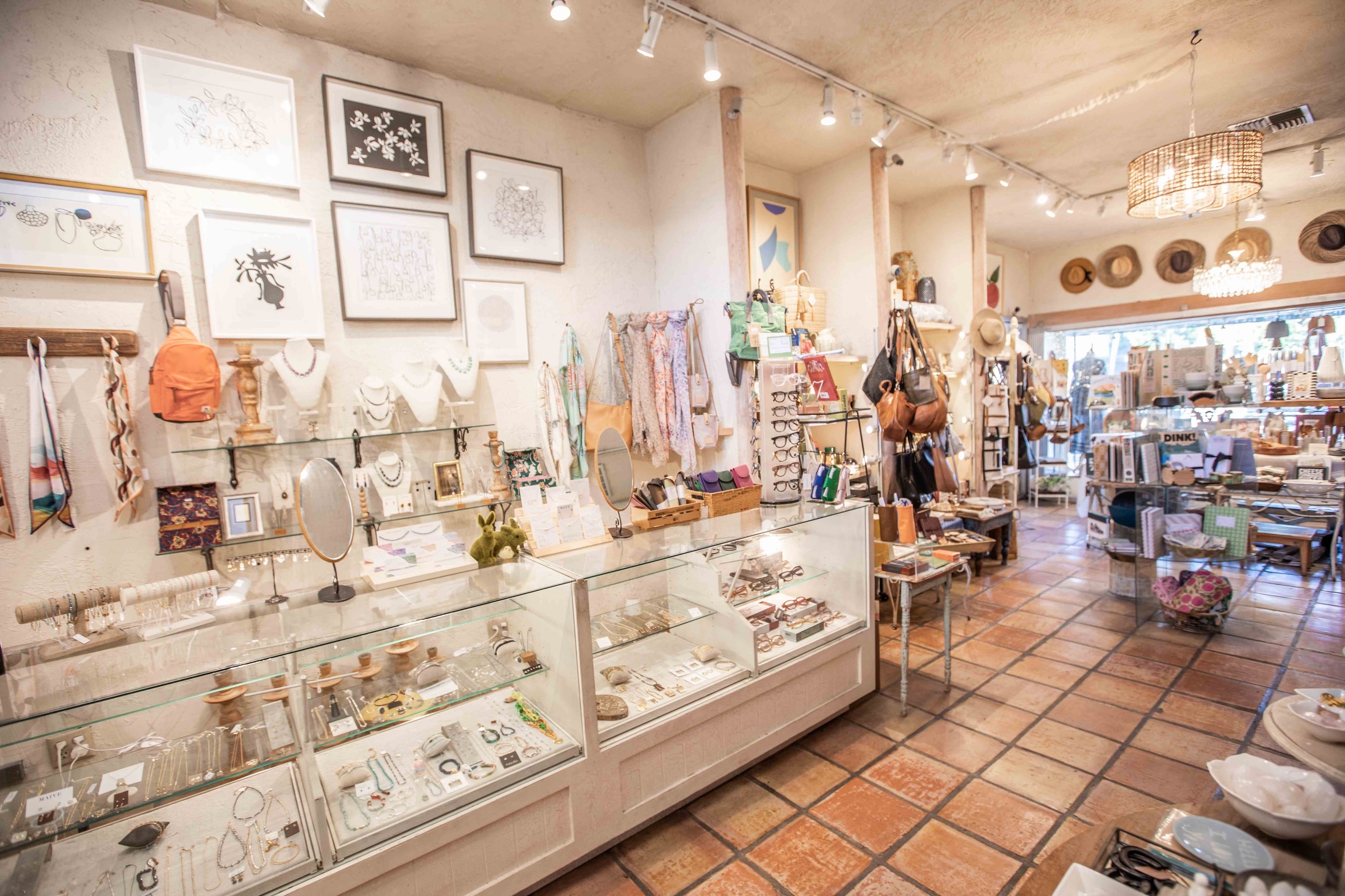 A cozy, well-lit boutique interior filled with jewelry display cases, framed wall art, hanging necklaces, and shelves of various items, set on terracotta tile floors.