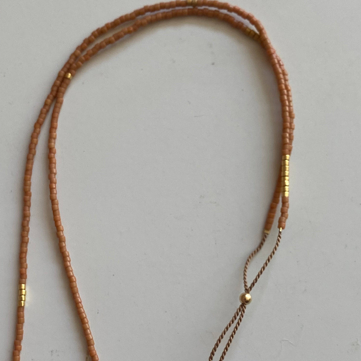 A long beaded necklace made of small warm brown beads with small gold spacer beads, laid out on a plain light gray surface. 