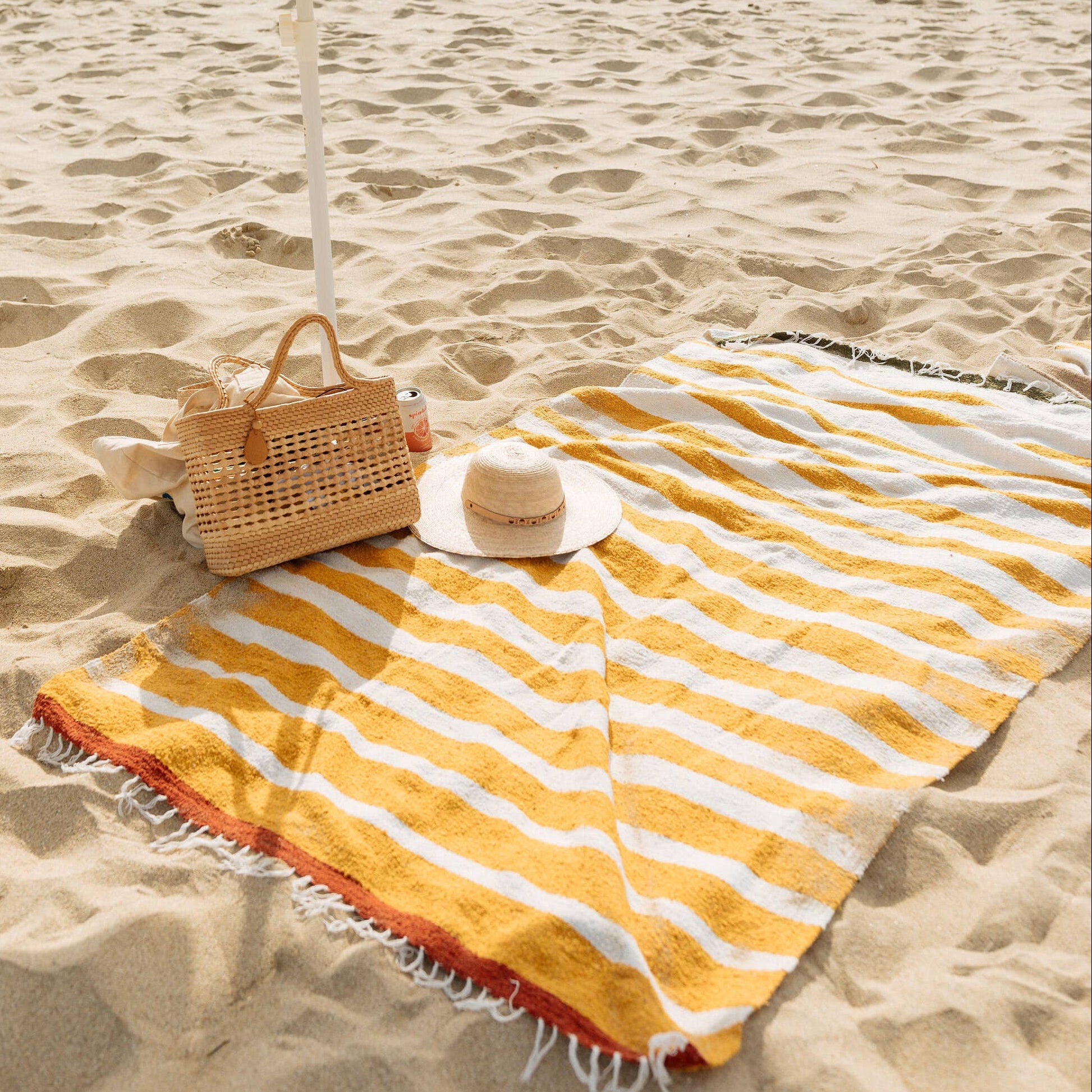 Yellow striped blanket, and woven bag on a sandy beach.