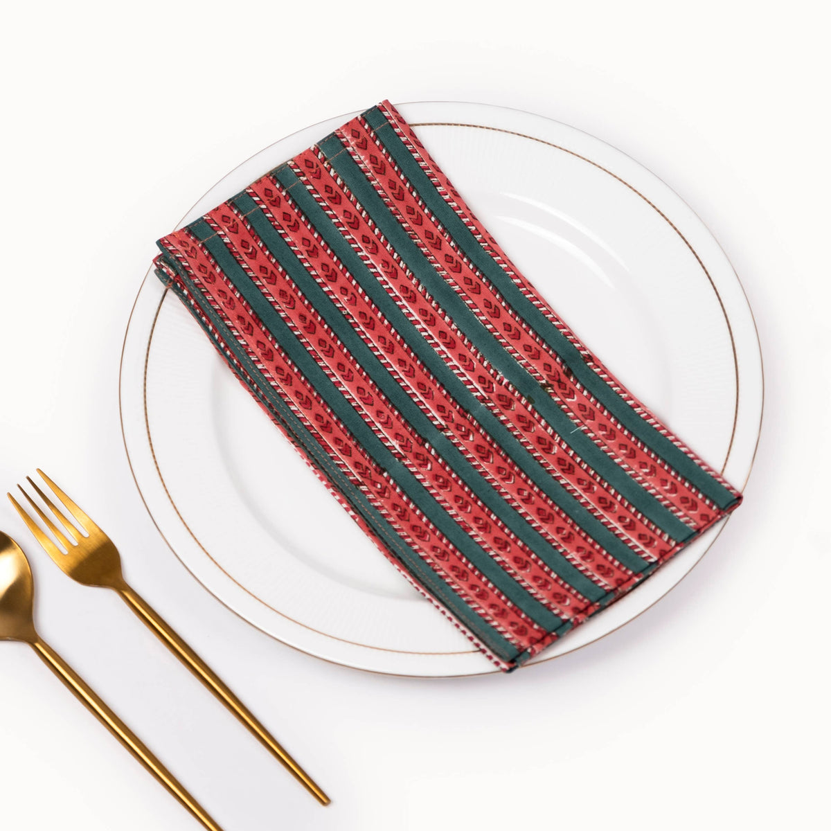 A folded striped napkin with red, green, and white patterns, placed on a white plate with gold trim, alongside gold-toned cutlery on a white background.