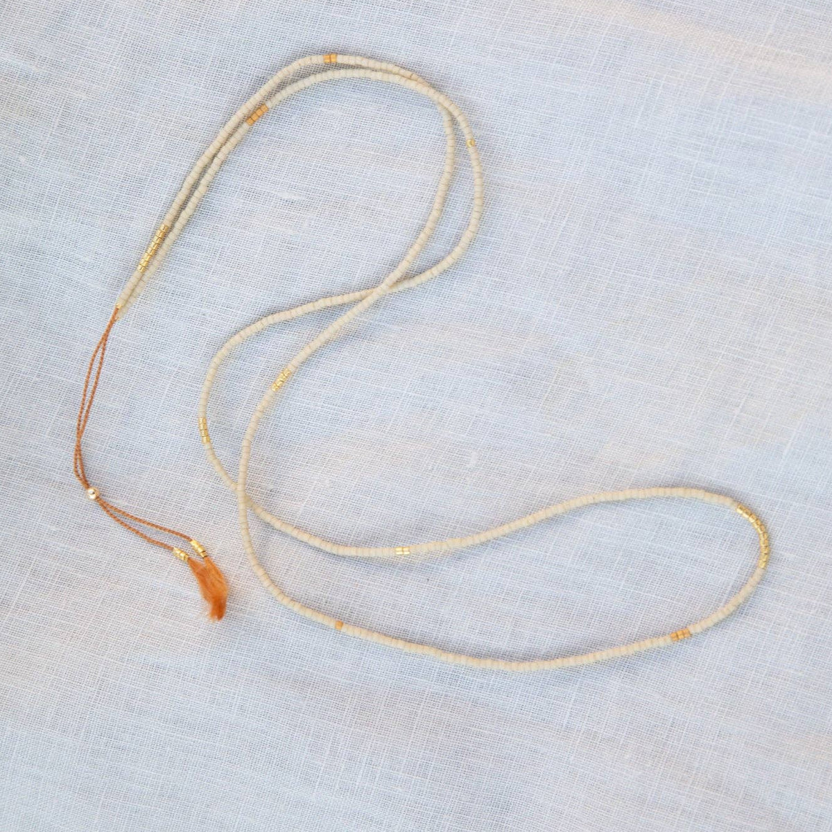 A delicate, thin rope-style necklace in a natural beige or light tan color, laid out on a textured, light gray fabric background.