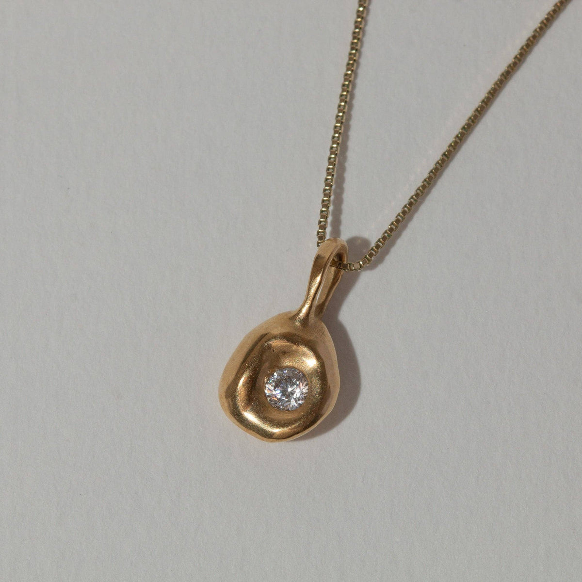 A gold pendant necklace with an organic, teardrop-shaped gold setting holding a small clear gemstone, shown on a plain light gray background.